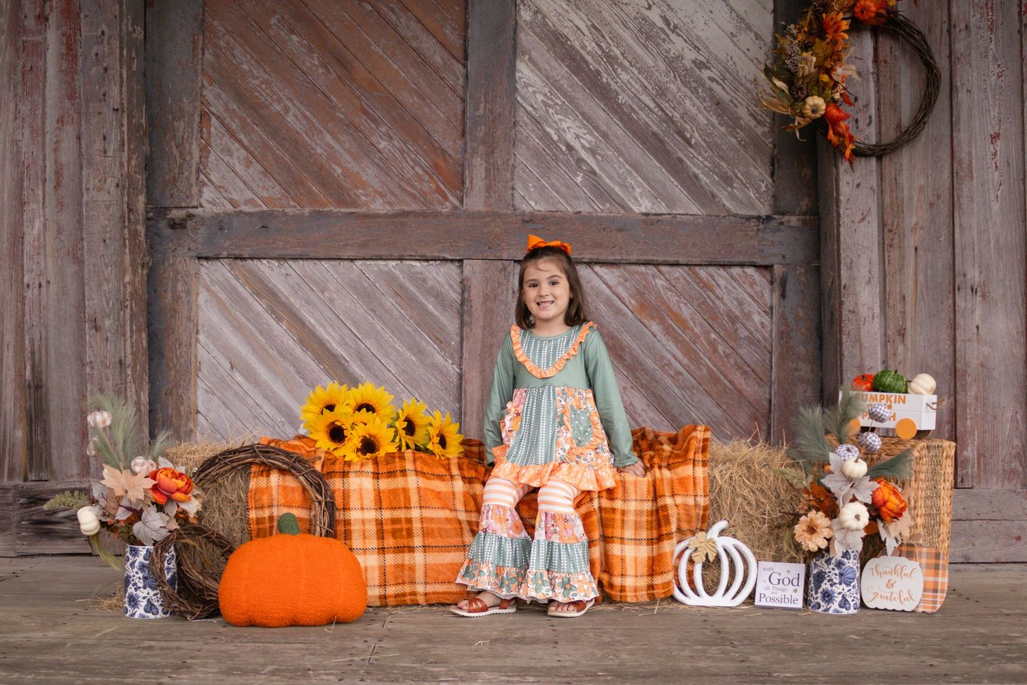 Young girl in teal and orange fall outfit poses with pumpkins and sunflowers by rustic barn doors.