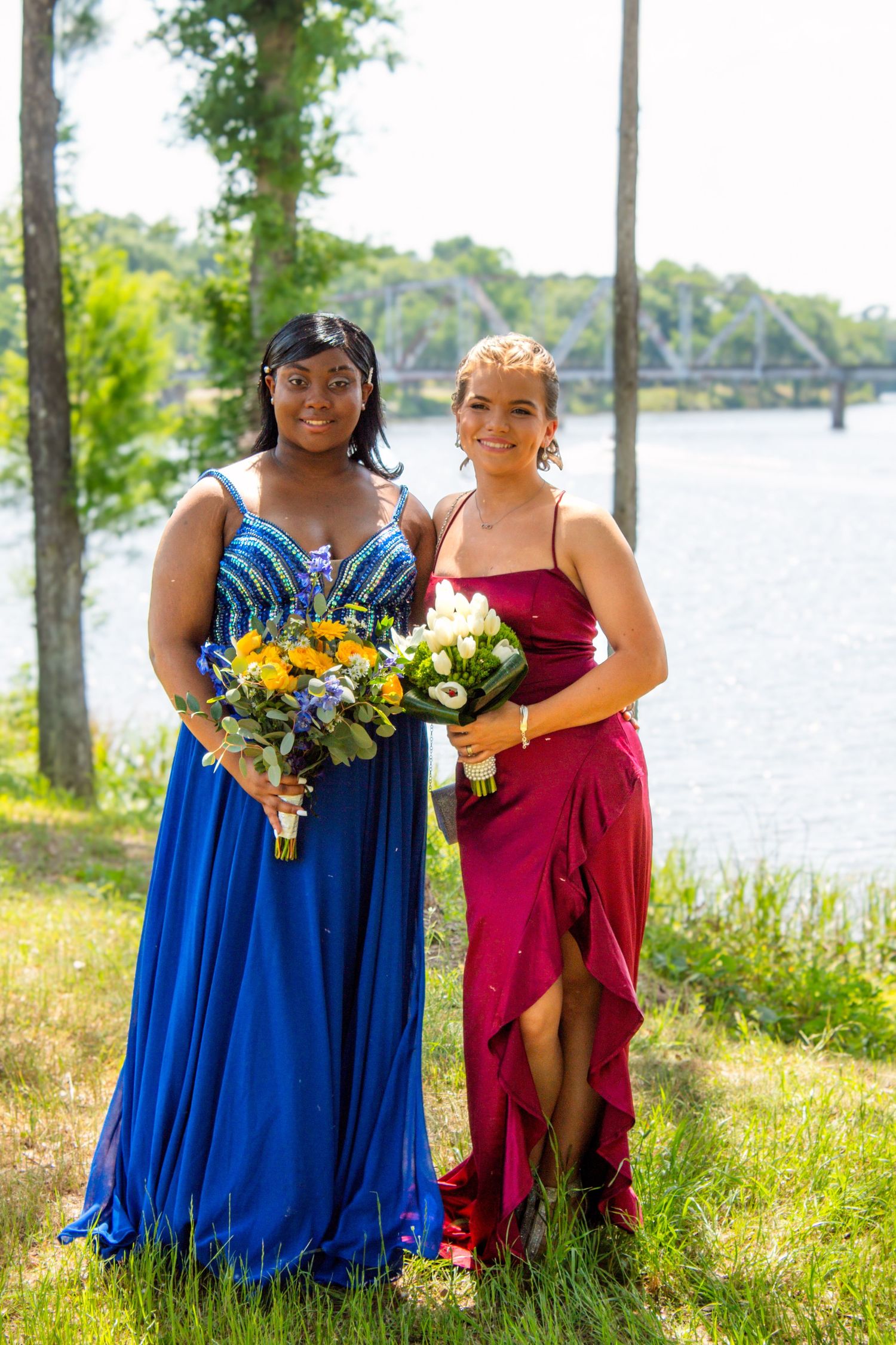 Two prom girls in blue and red gowns hold bouquets while posing by a sunny riverside.