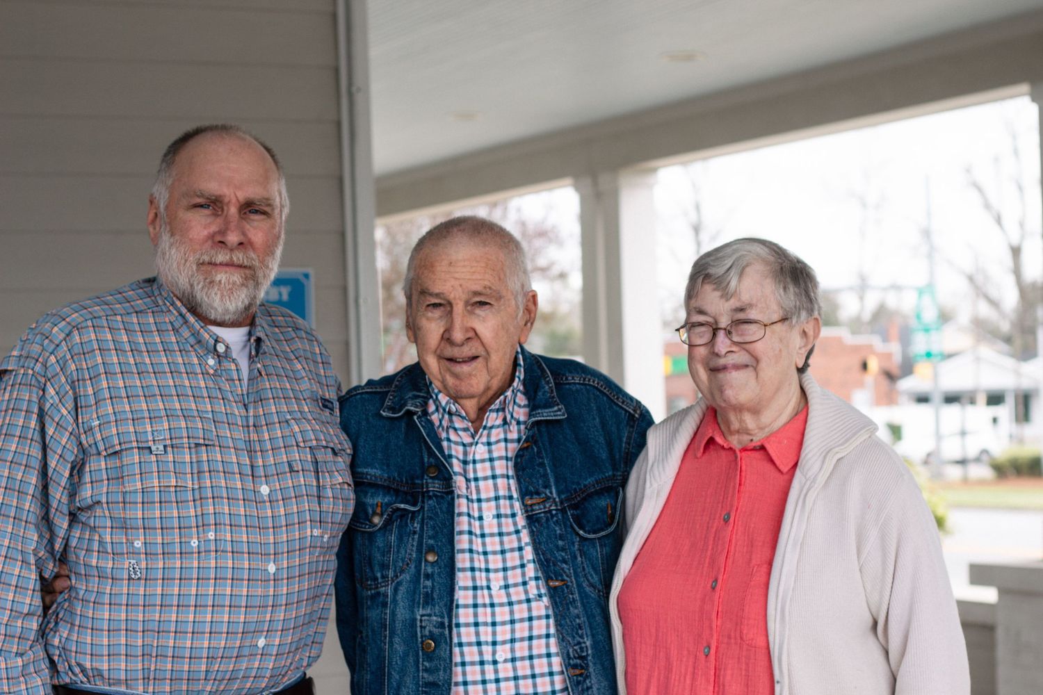 Three elderly adults stand together smiling on a covered porch in a casual family photo.