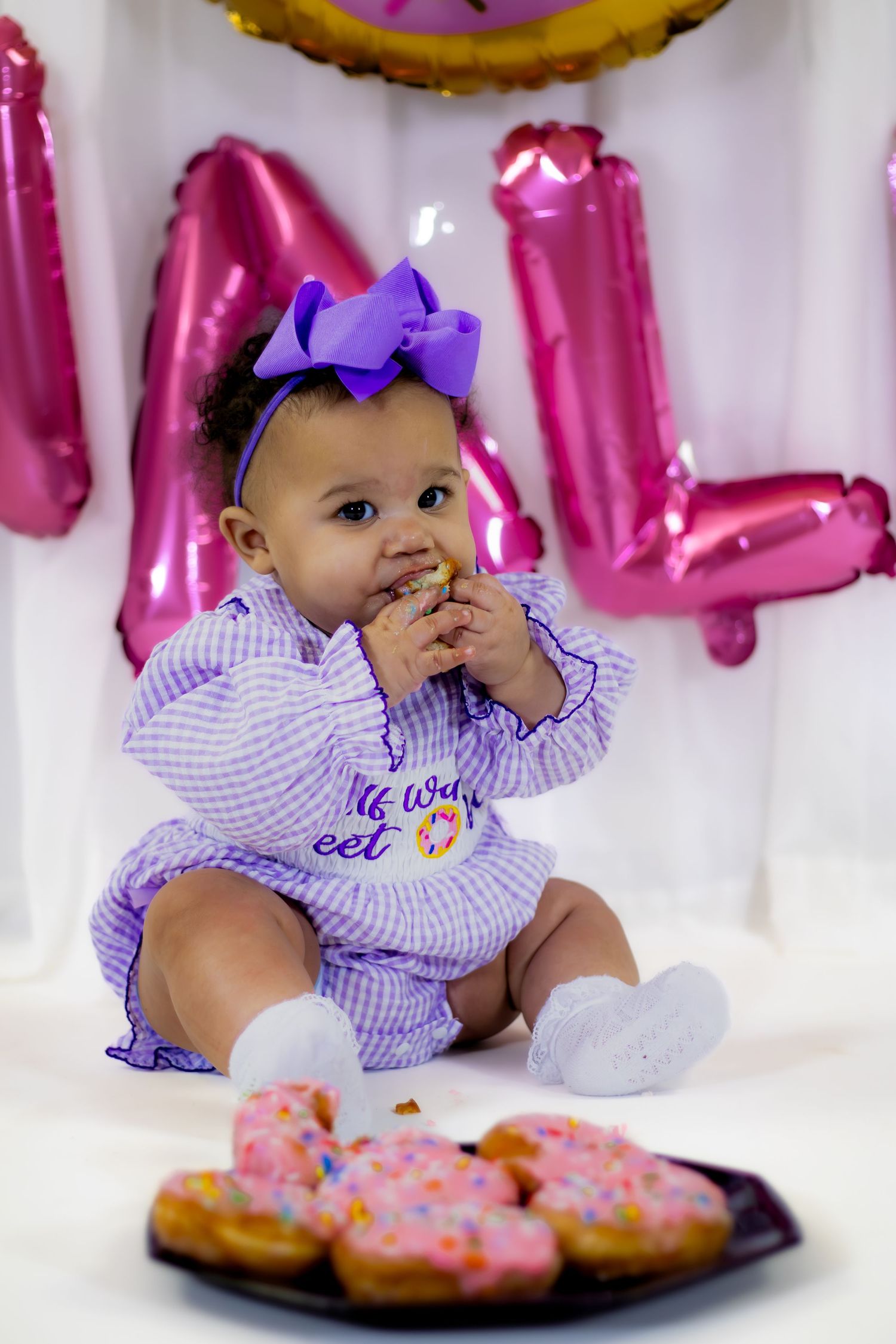 Baby girl in purple bow headband eating donut at colorful half birthday party with pink balloons.