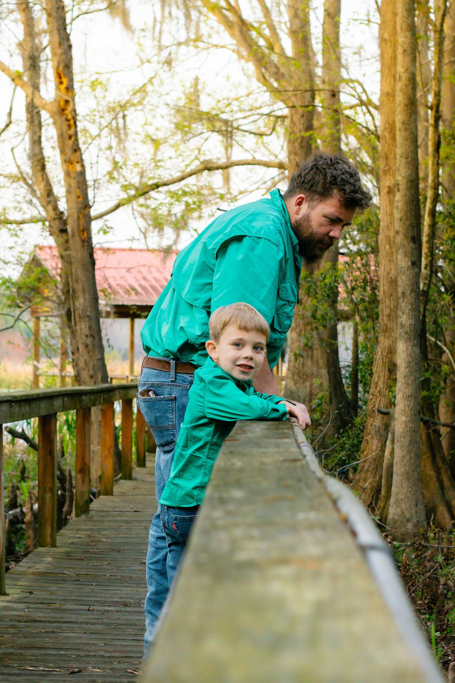 Father and young son in green shirts lean on wooden bridge railing surrounded by trees.