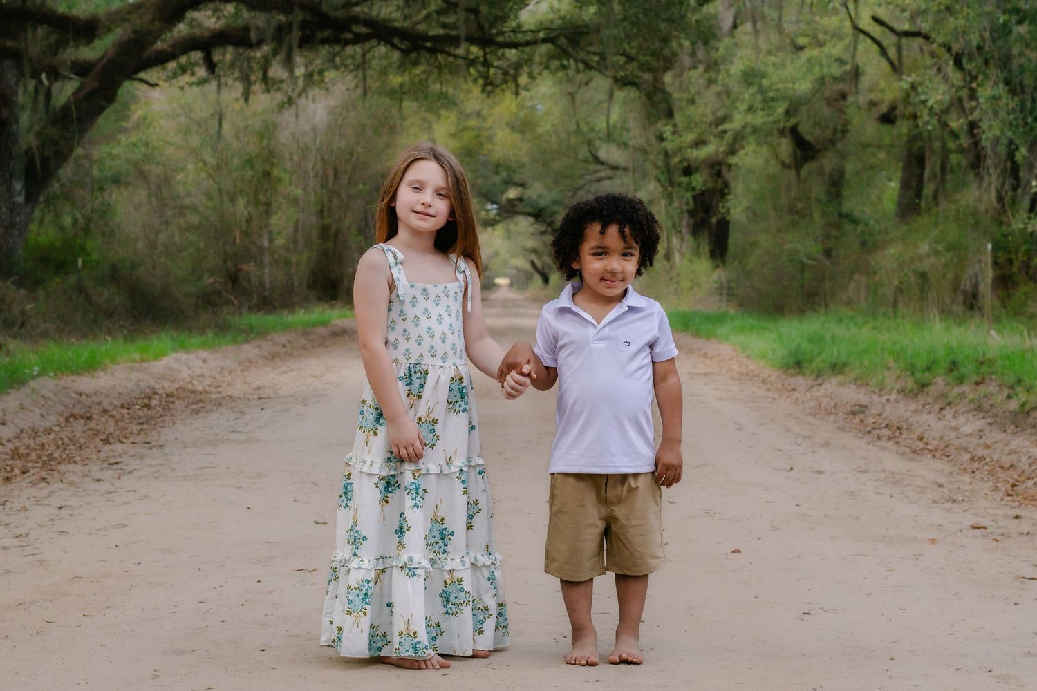 A girl and boy in summer outfits hold hands on a tree-lined dirt path.