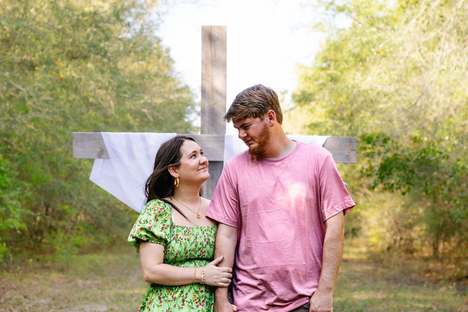 Couple gazing at each other lovingly outdoors with a wooden cross in the background surrounded by trees.