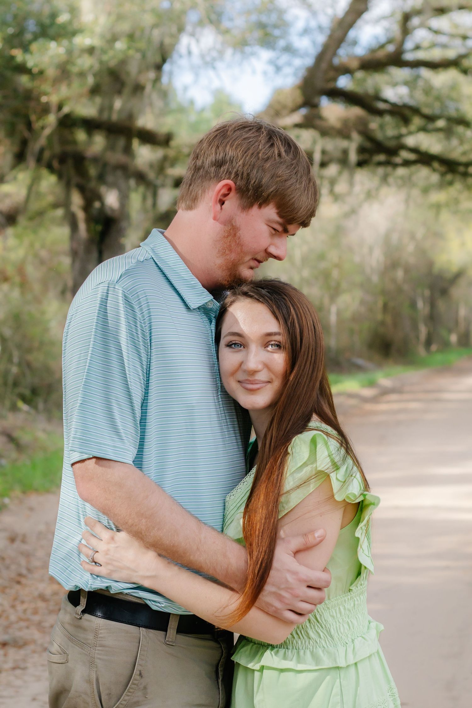 Couple embracing tenderly on a tree-lined path, woman in green dress smiling at camera.