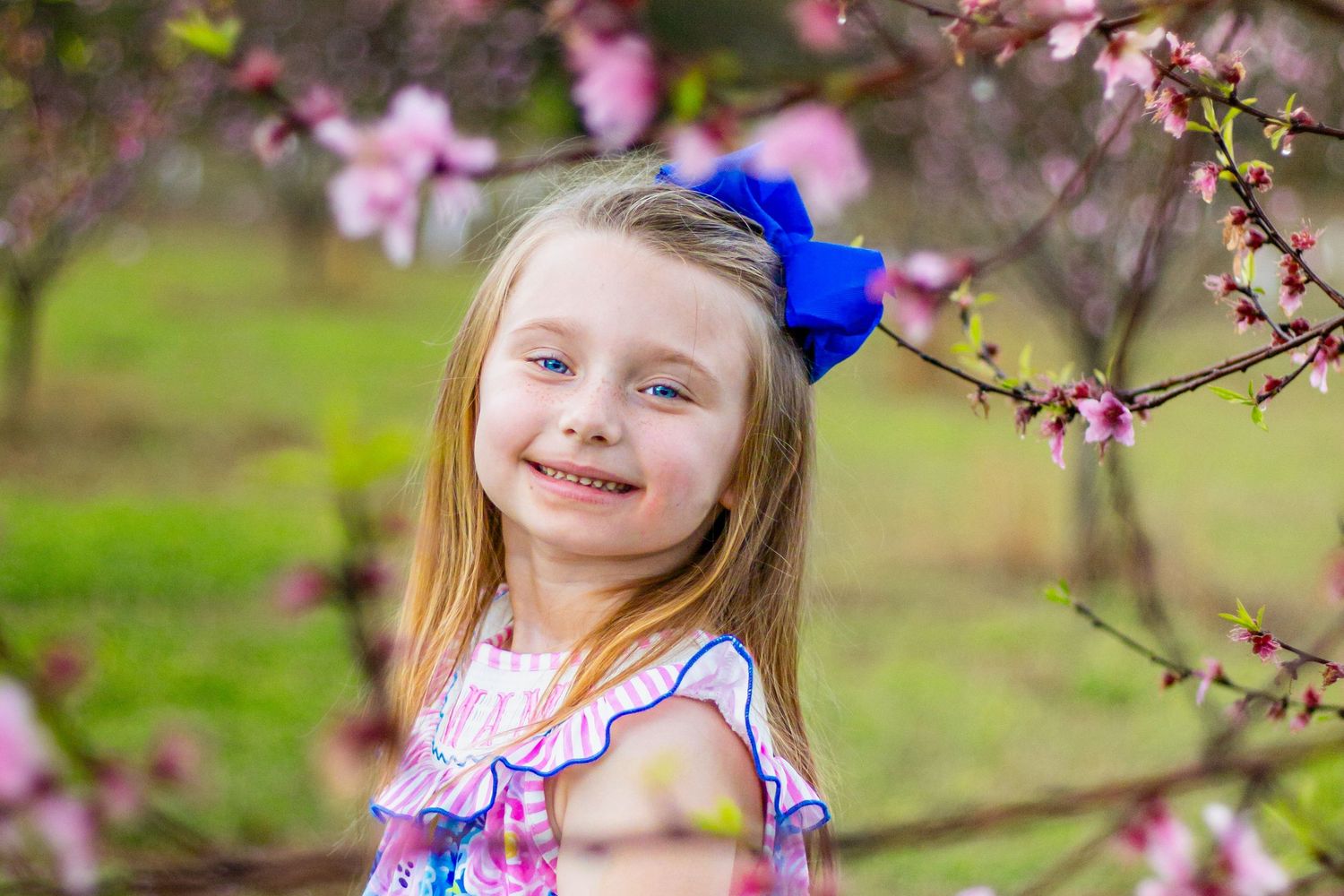 Happy blonde girl with blue hair bow smiles beside pink flowering tree branches in a spring garden setting.