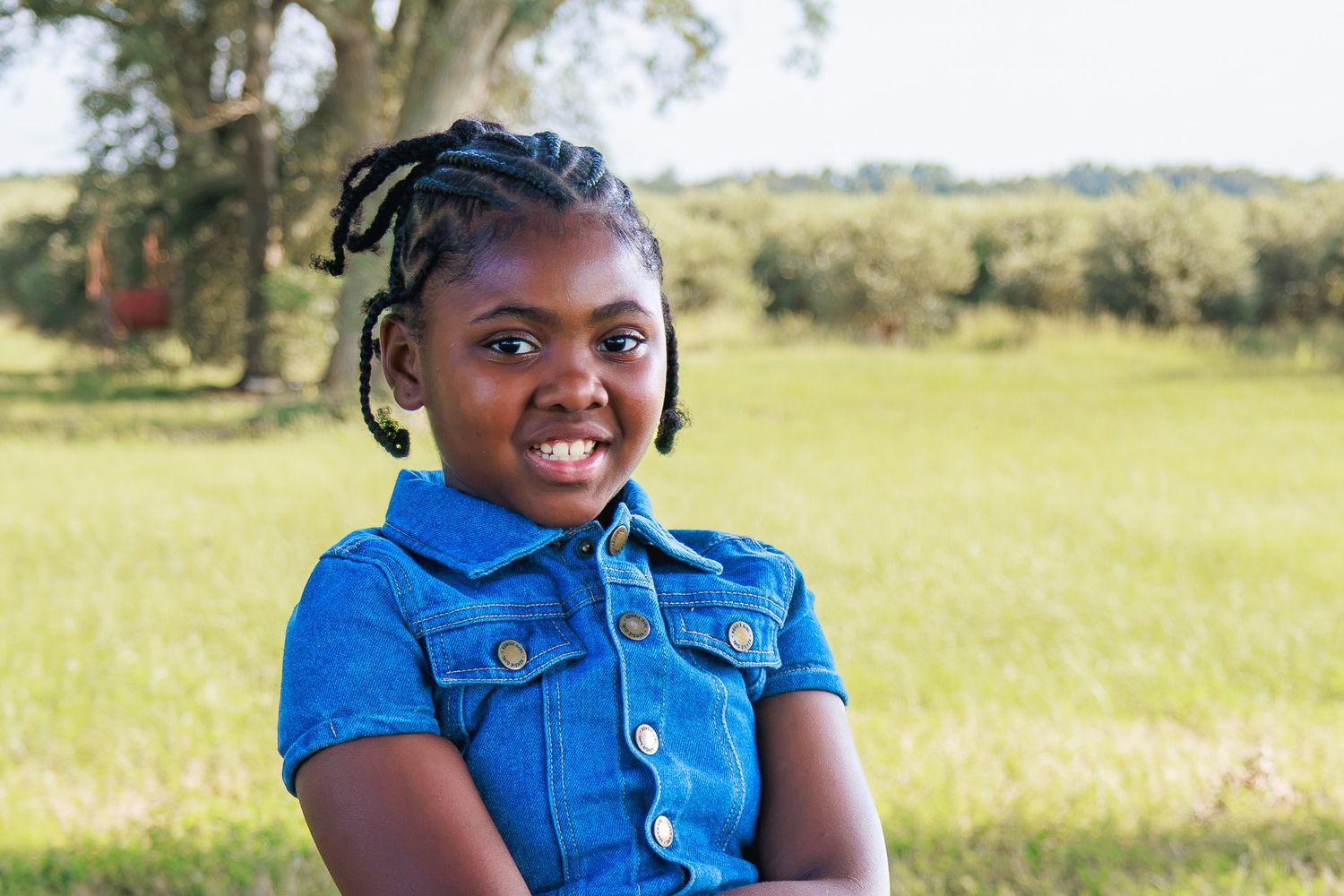 A smiling young African girl with braids in a blue denim vest poses outdoors in a green field.
