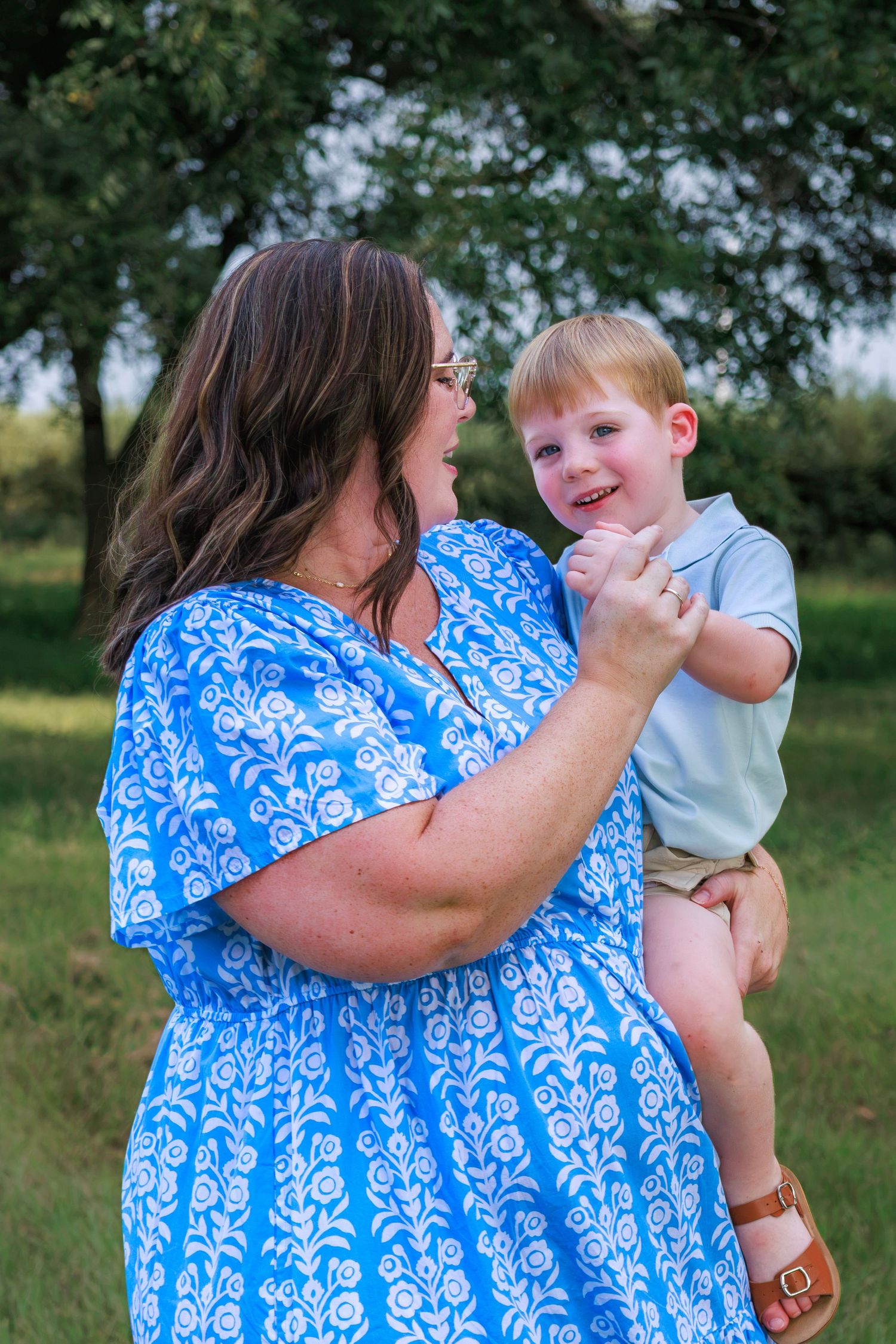 A woman in a blue floral dress holds a smiling toddler boy in a sunny outdoor park setting.