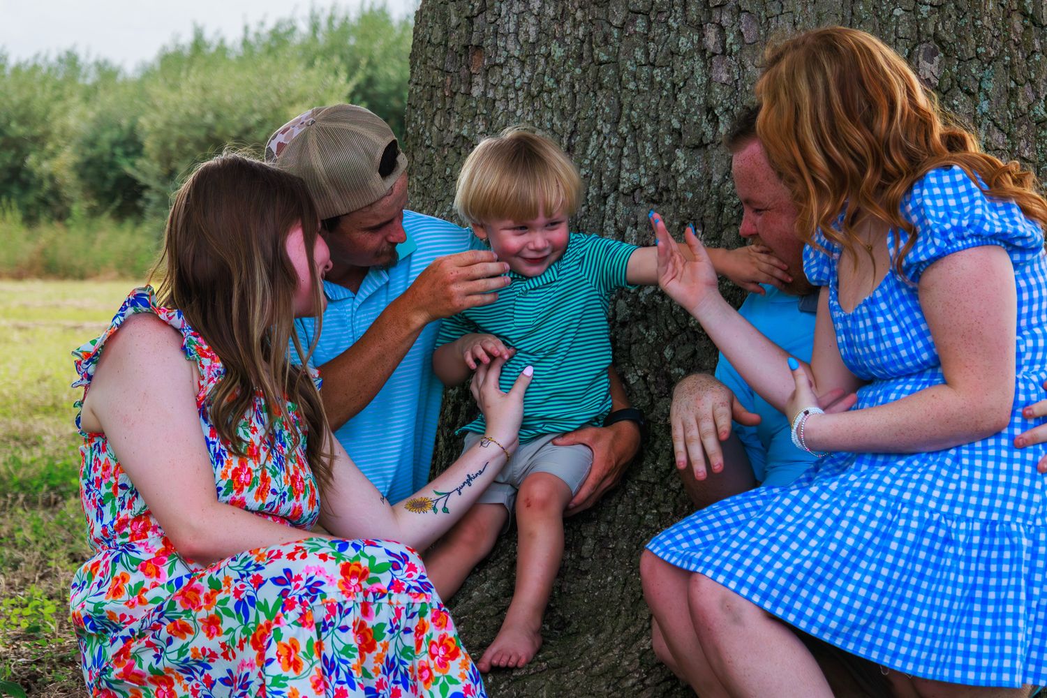 Family of four laughing together by a tree outdoors, with children and parents in colorful summer outfits.