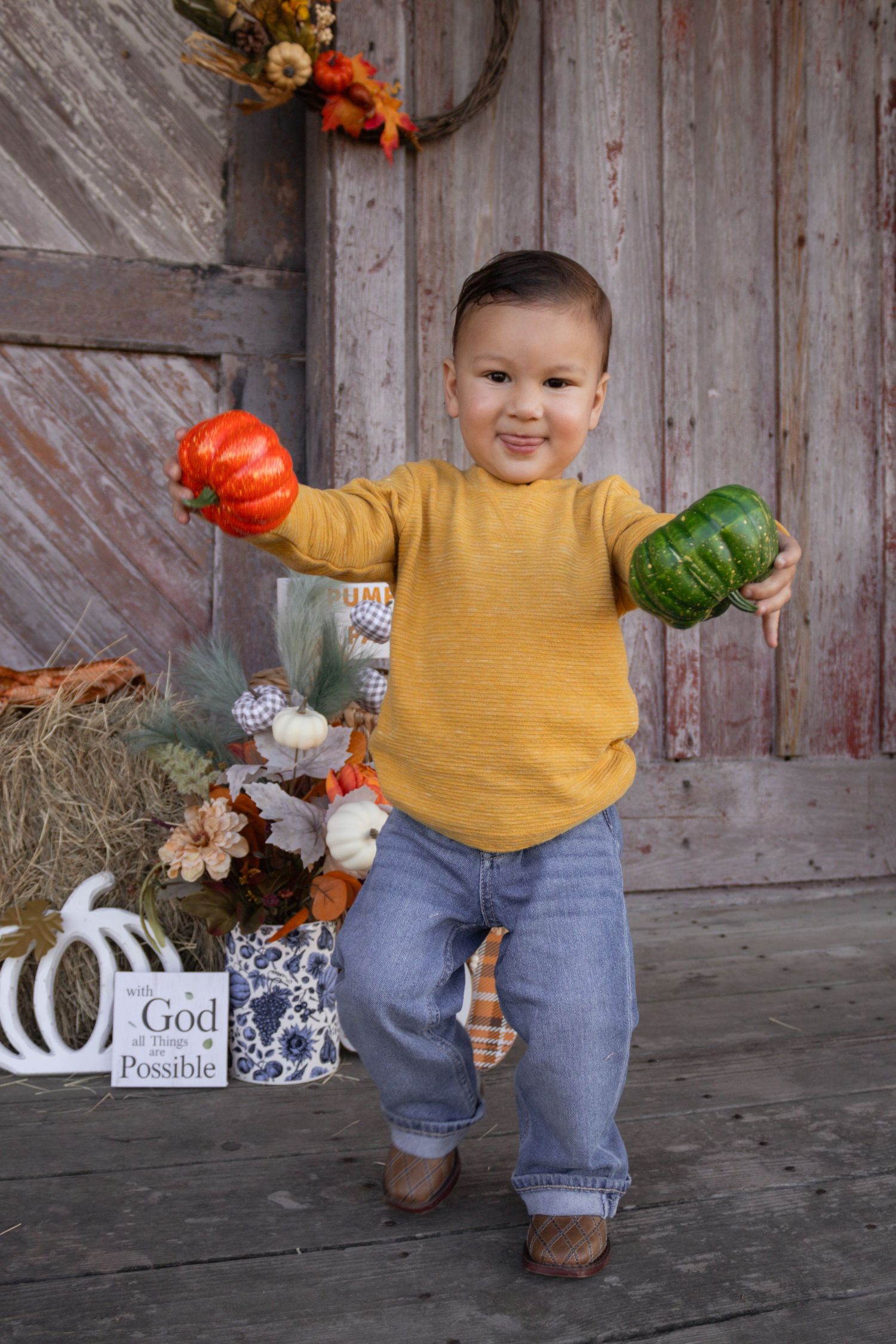 Cheerful toddler boy in yellow sweater holding orange and green gourds at fall barn backdrop.