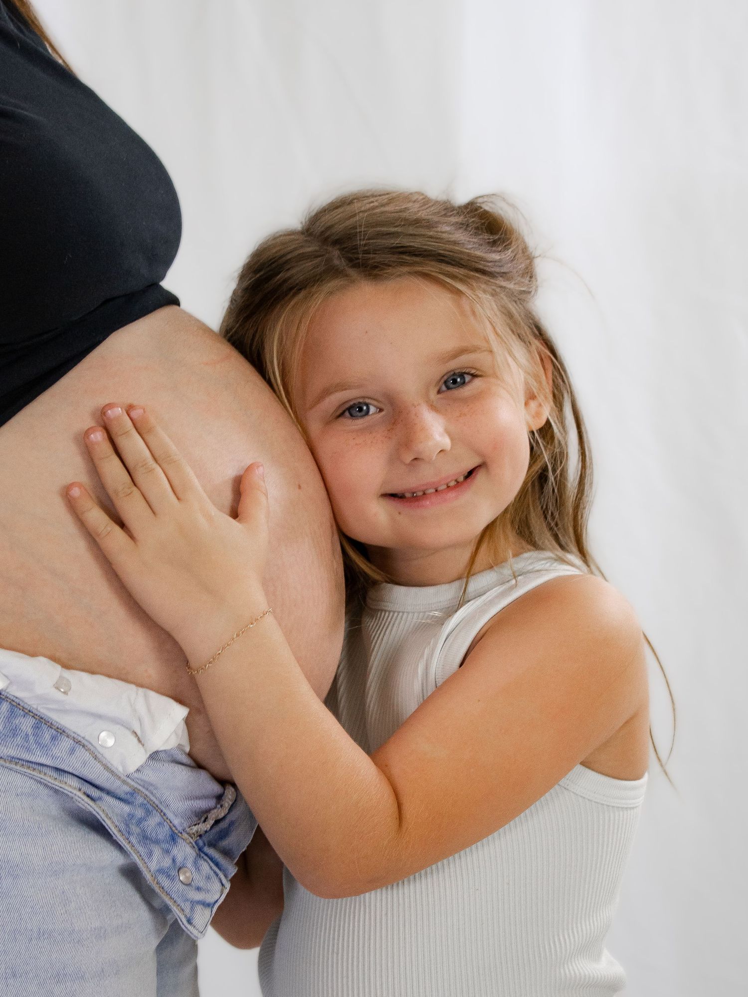 Smiling young girl hugging and pressing her cheek against a pregnant womans baby bump indoors.
