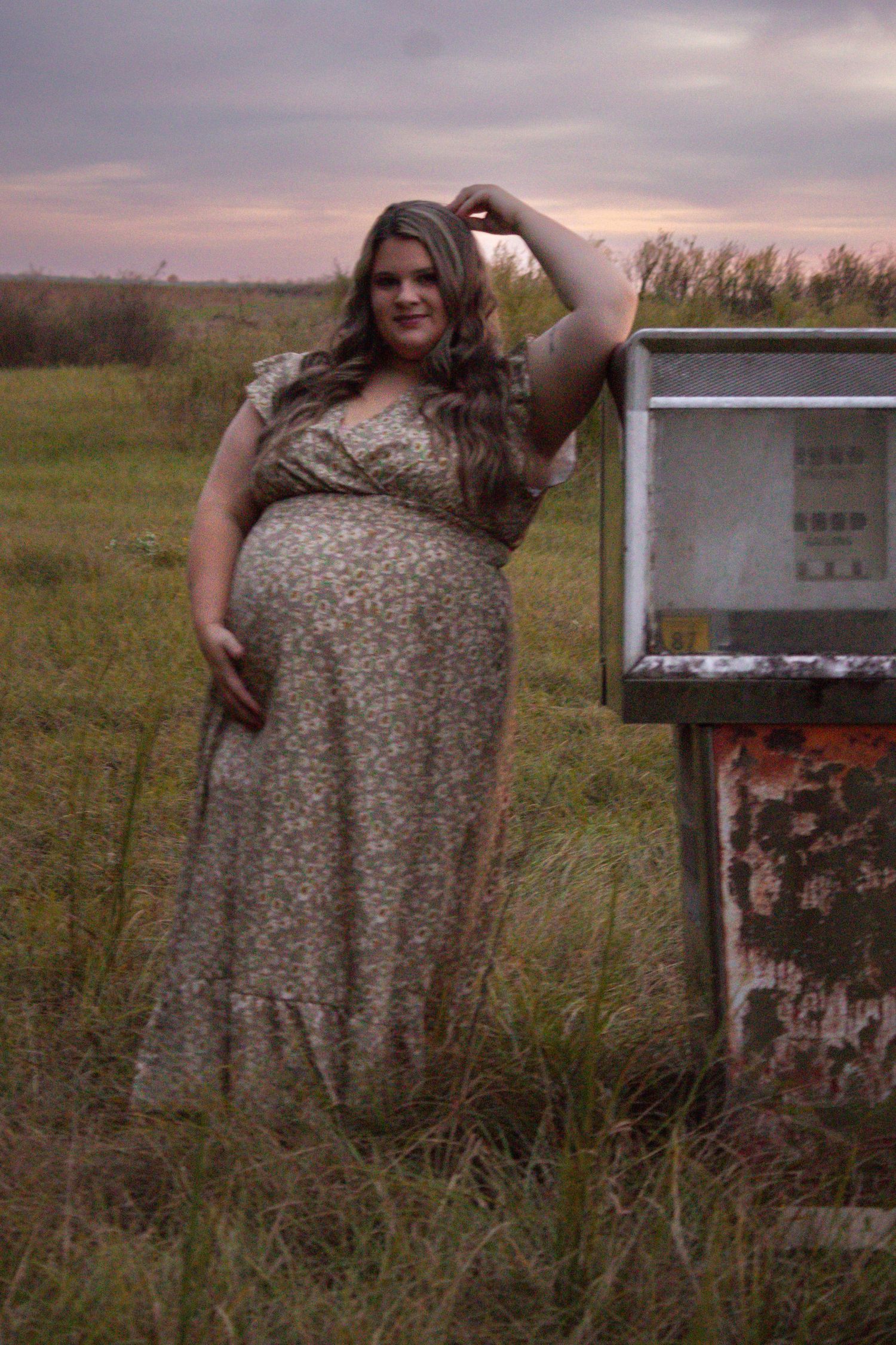 Pregnant woman in floral dress posing by rustic box in grassy field at dusk.