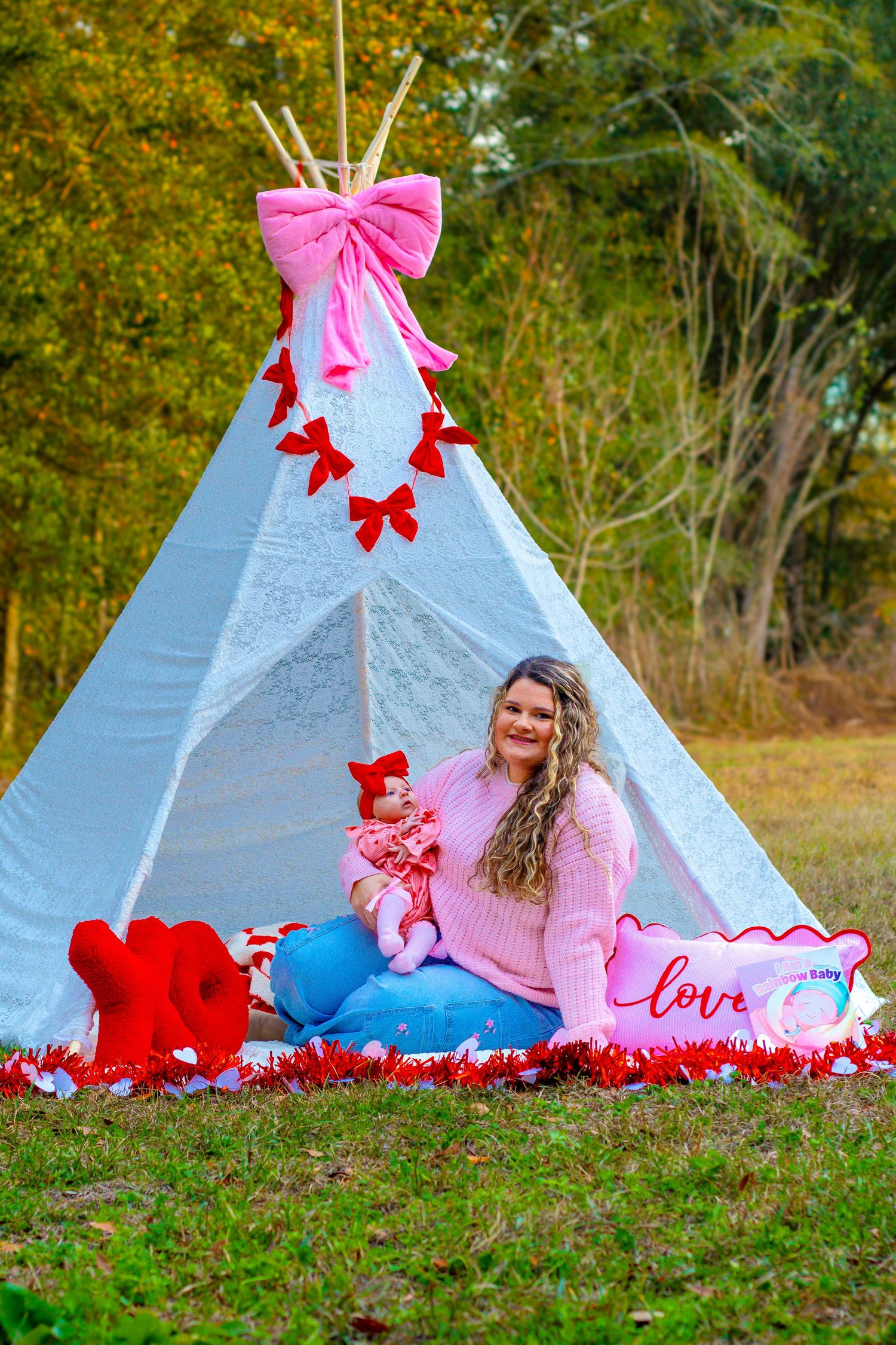 Woman in pink sweater holds baby in red bow near decorated white teepee with pink bows and Valentine props.
