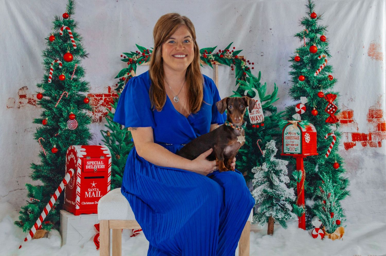 A smiling woman in a blue dress holds a dachshund while seated in a Christmas-decorated studio.