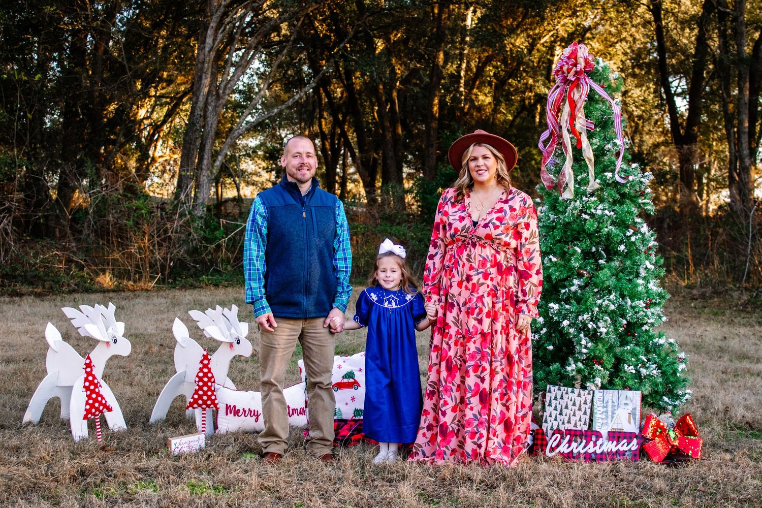 A family of three poses outdoors with white reindeer decorations and a festive Christmas tree.