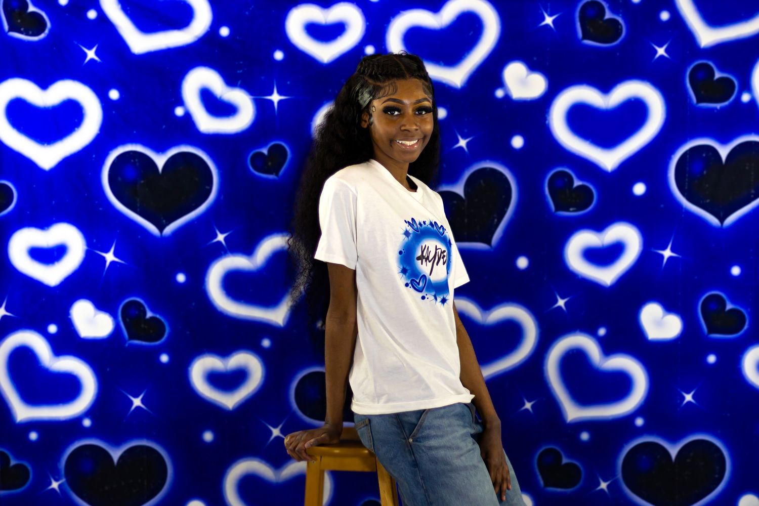 Young woman sitting on a stool smiling in a white t-shirt against a blue heart backdrop.