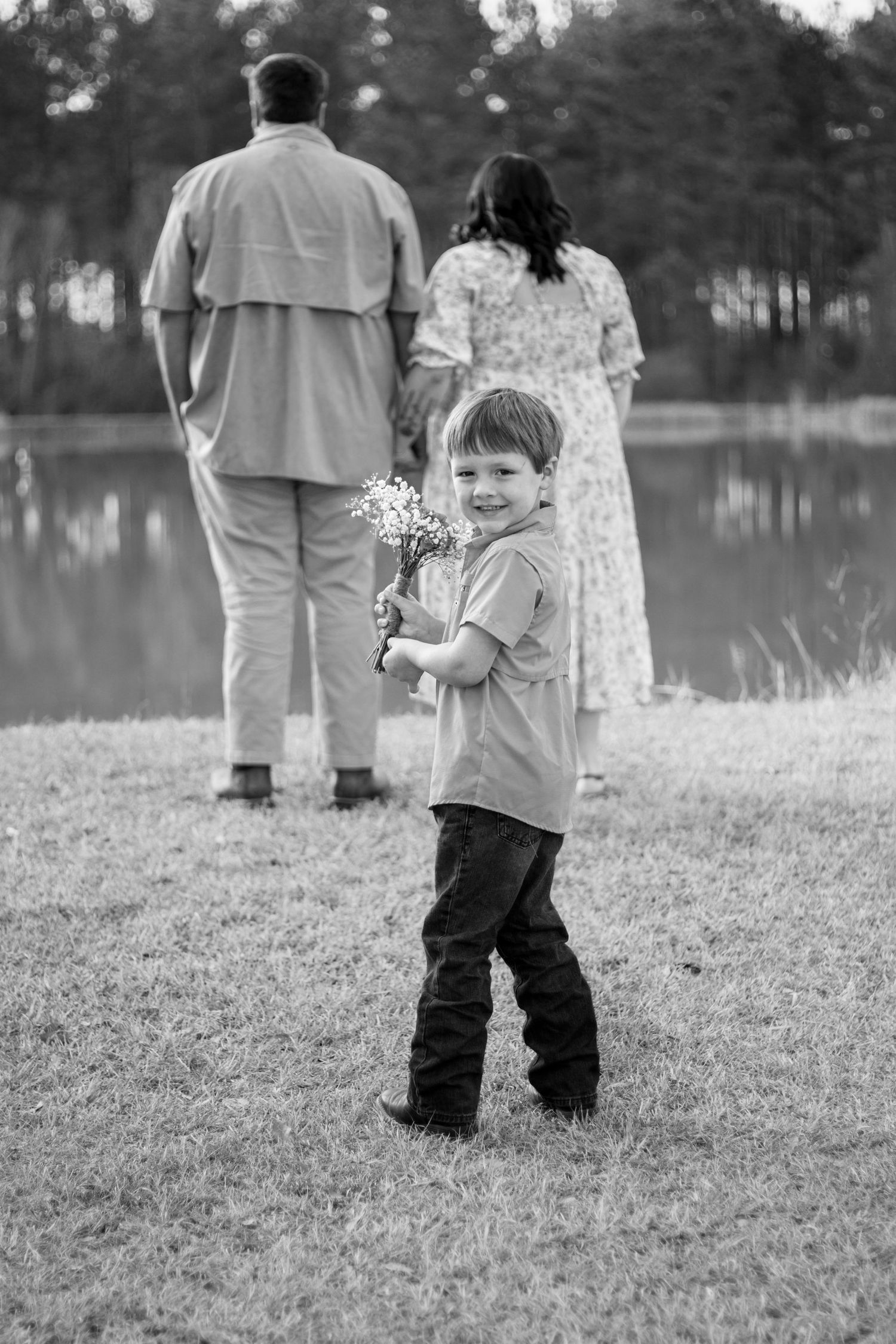 A young boy holds wildflowers and smiles at the camera while his parents stand by a serene pond in black and white.