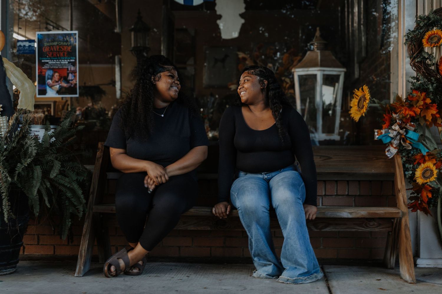 Two young Black women in black outfits sit on a bench outside a decorated storefront.