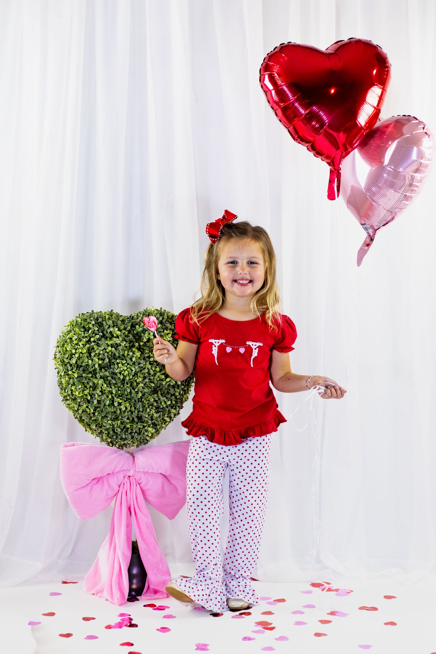 Smiling girl in red top and polka dot pants holds heart balloons near a heart-shaped topiary with pink bow.