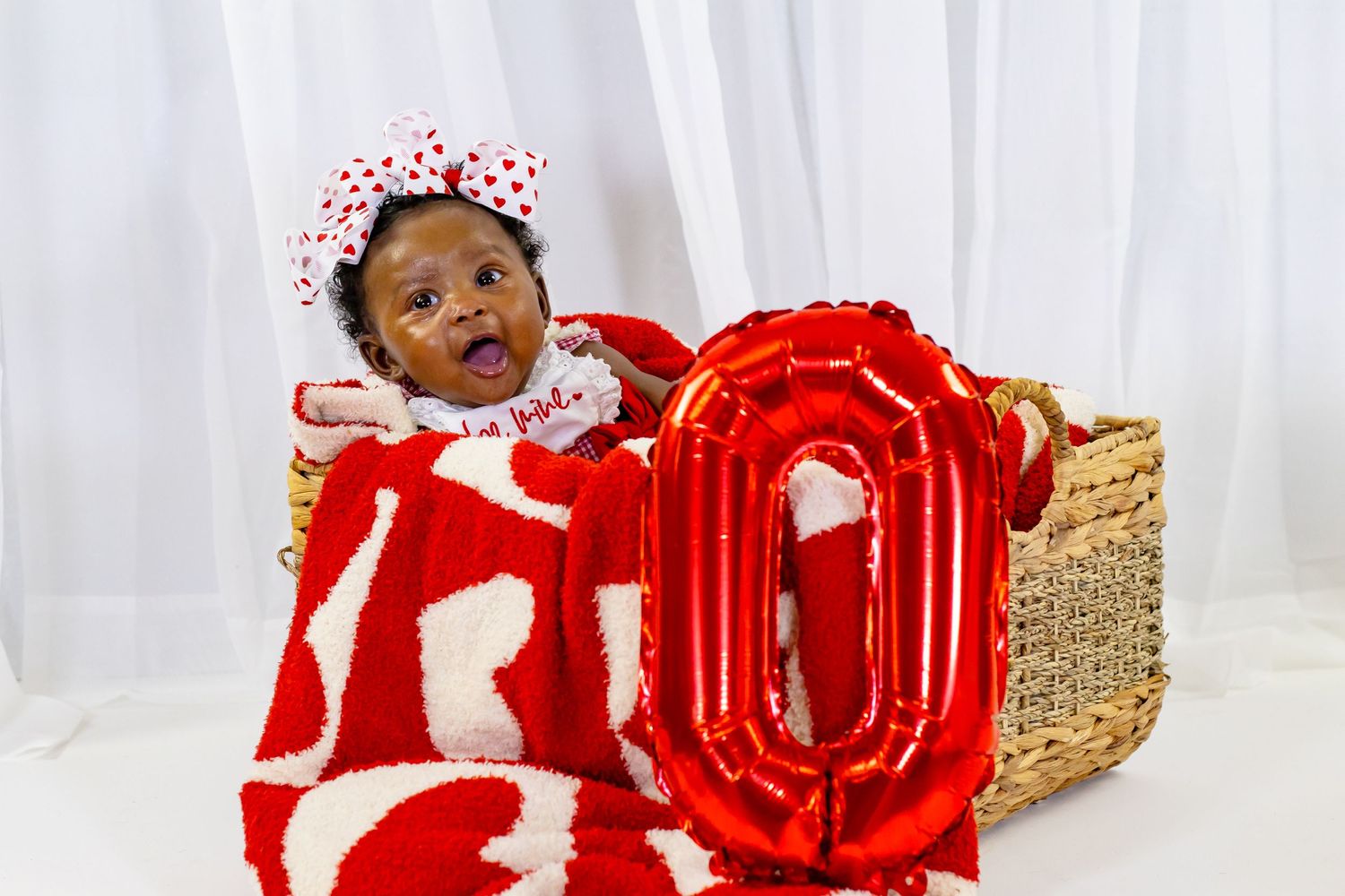 Baby girl wrapped in red blanket sitting in basket with red balloon number zero for Valentine's Day photo.