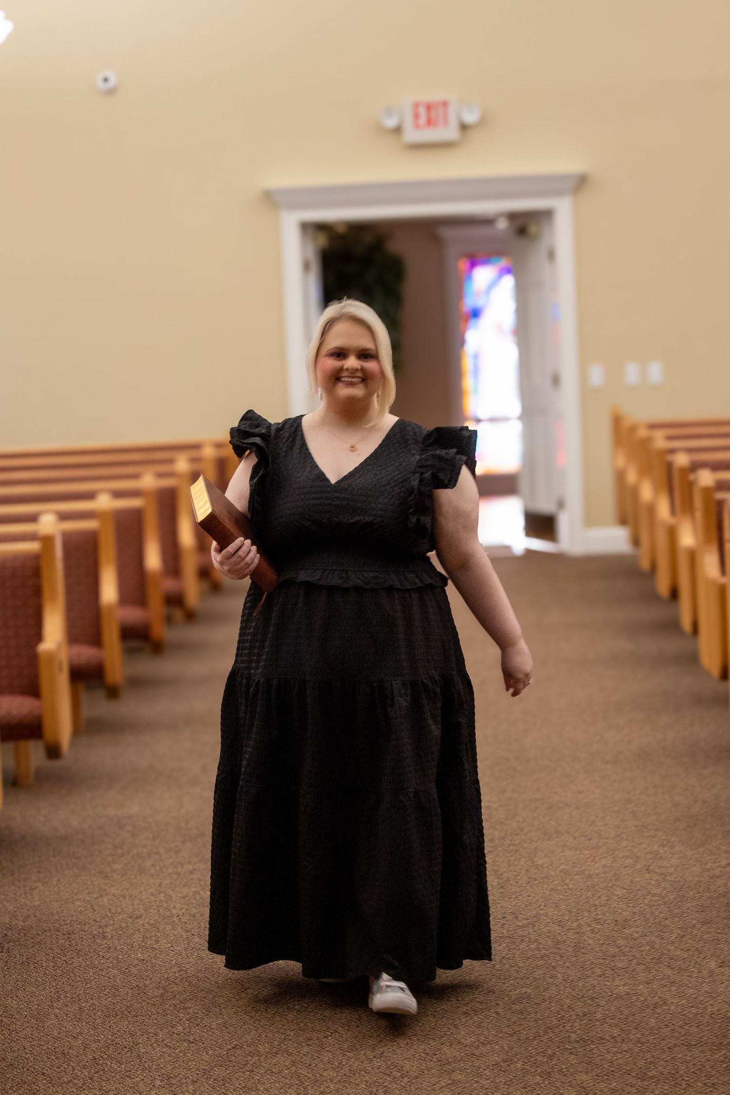 Blonde woman in black maxi dress walking down church aisle holding a Bible toward camera.
