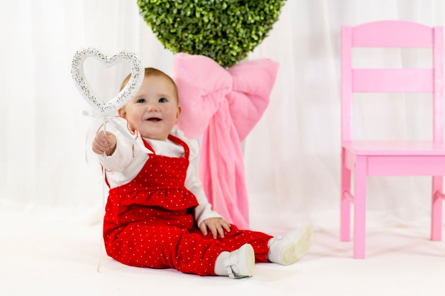Happy baby in red polka dot overalls holds a heart wand next to a pink chair for Valentine's Day photos.