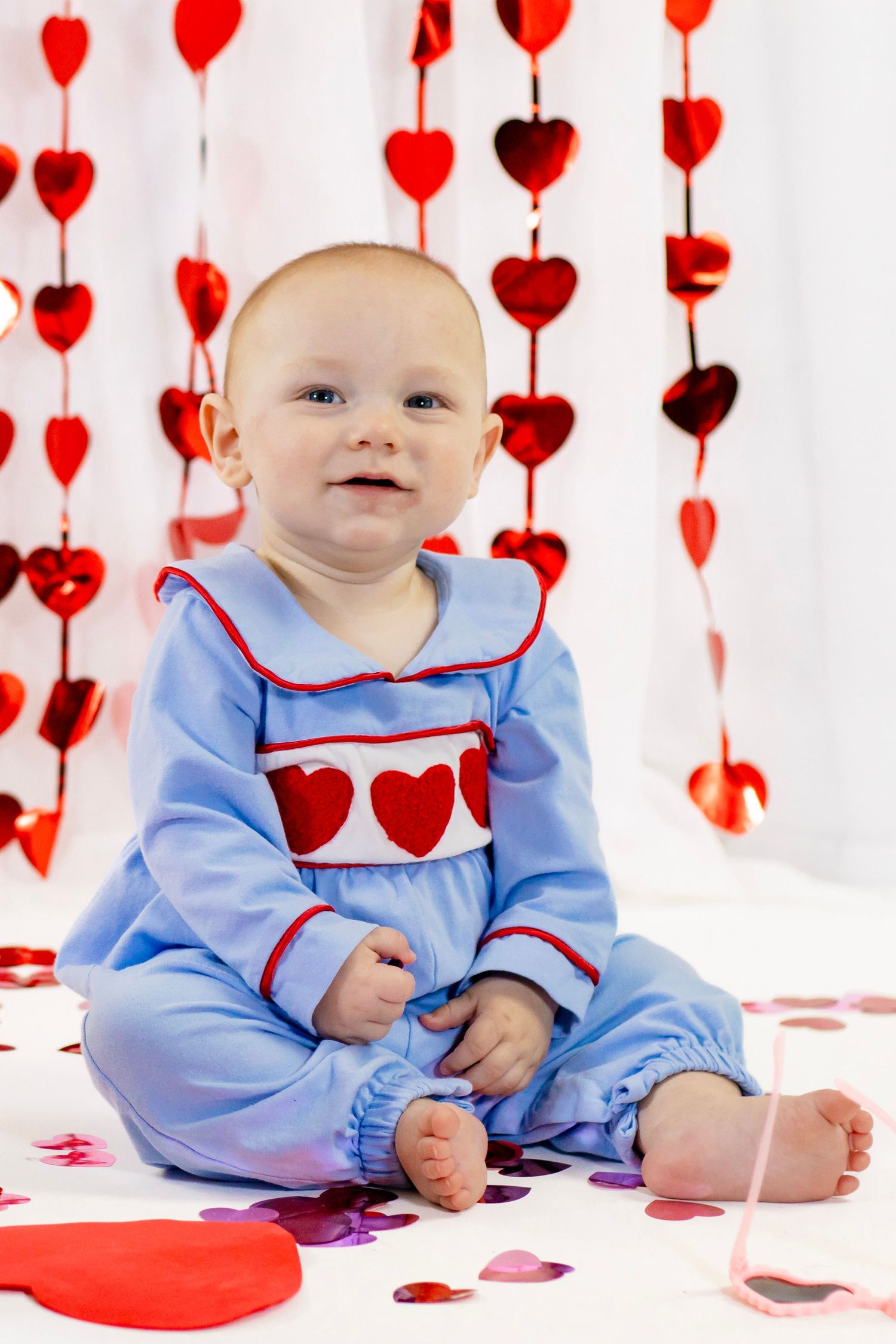 Smiling baby in blue heart romper sits among red heart garlands and confetti for Valentine's Day.