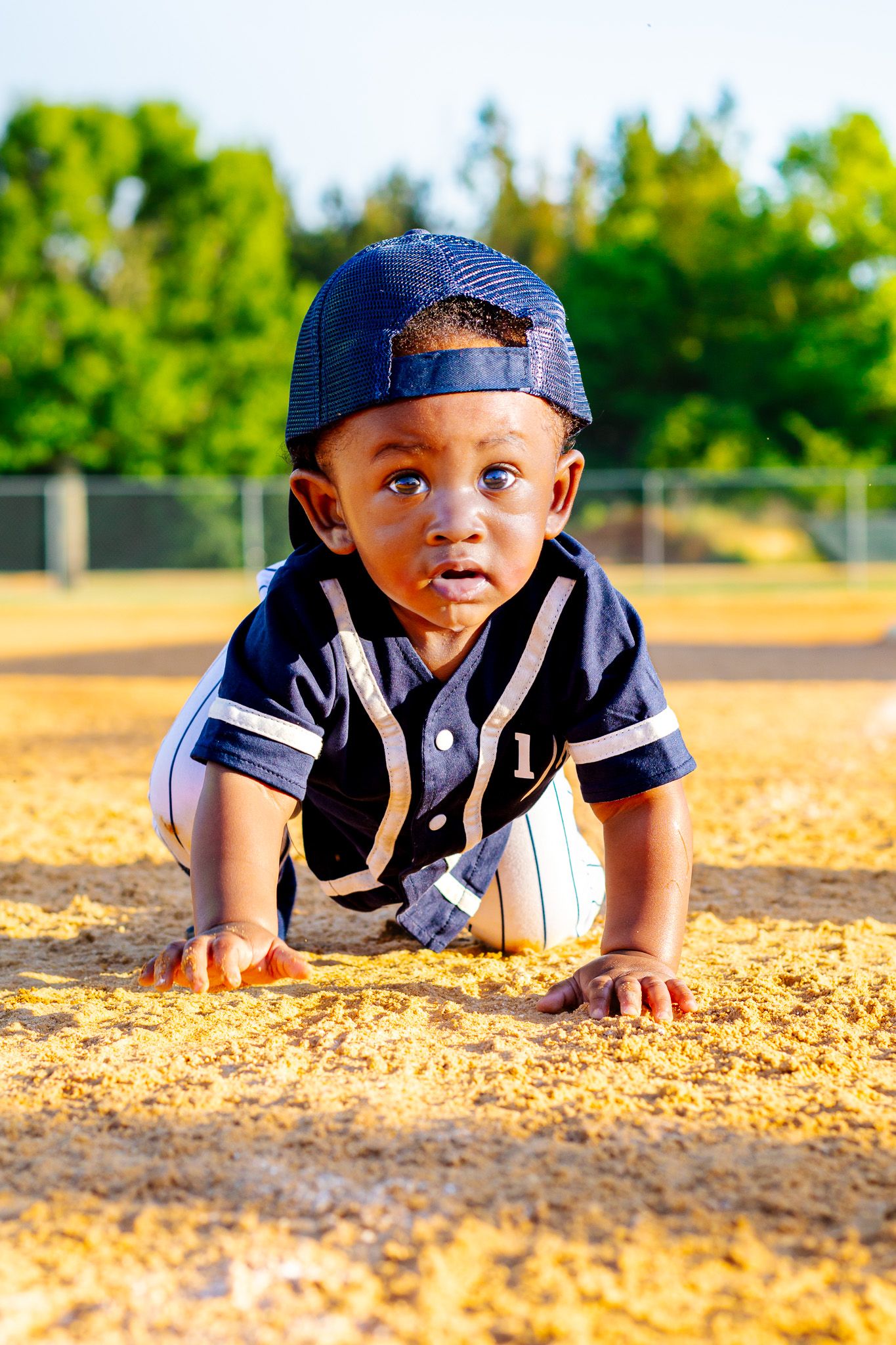 Adorable toddler in a baseball uniform crawling on a sandy baseball diamond field outdoors.