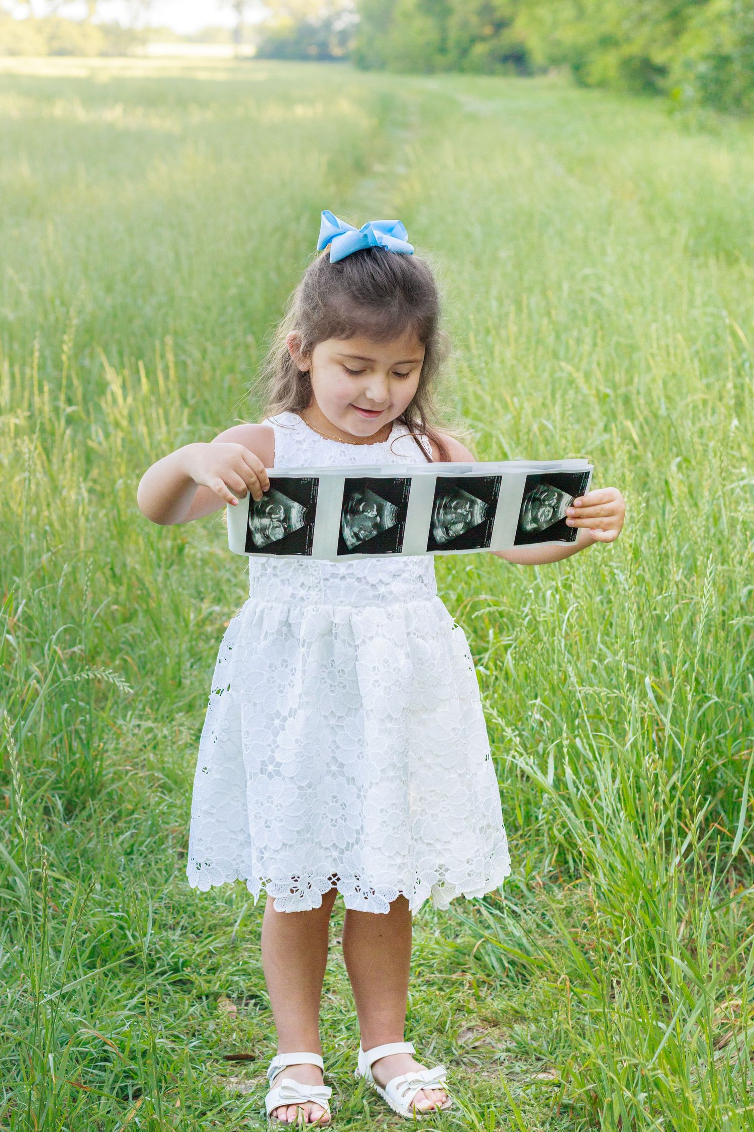 Young girl in white dress holding ultrasound photos in a green field, announcing a new sibling.