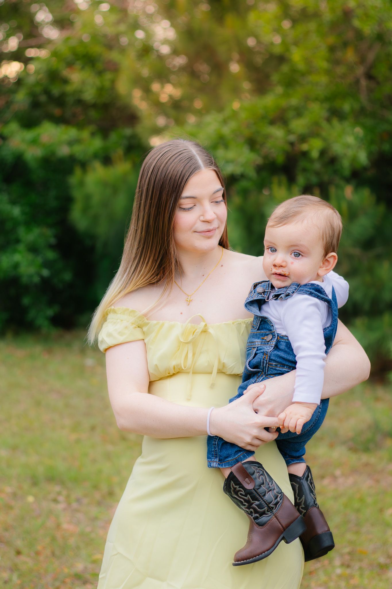 A teenage girl in a yellow dress lovingly holds a baby dressed in denim overalls and cowboy boots outdoors.