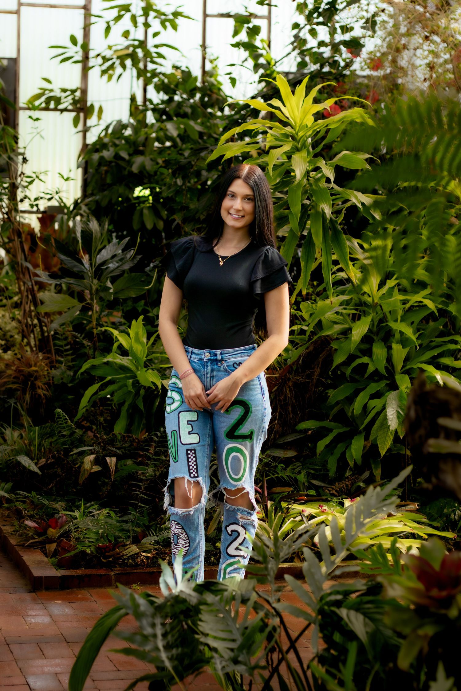 Young woman in black top and painted ripped jeans smiles in lush greenhouse surrounded by tropical plants.