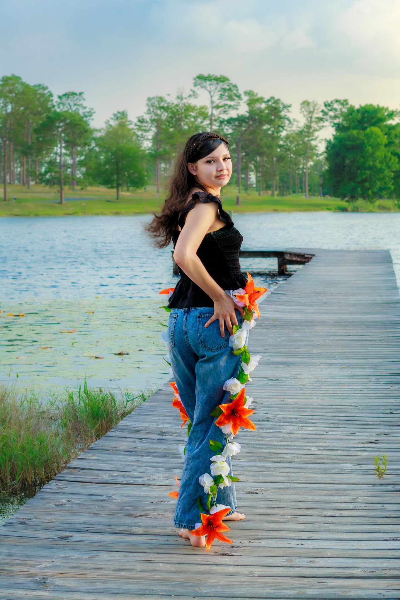 Young woman in jeans and black top poses on wooden dock by serene lake, adorned with colorful flower garland.