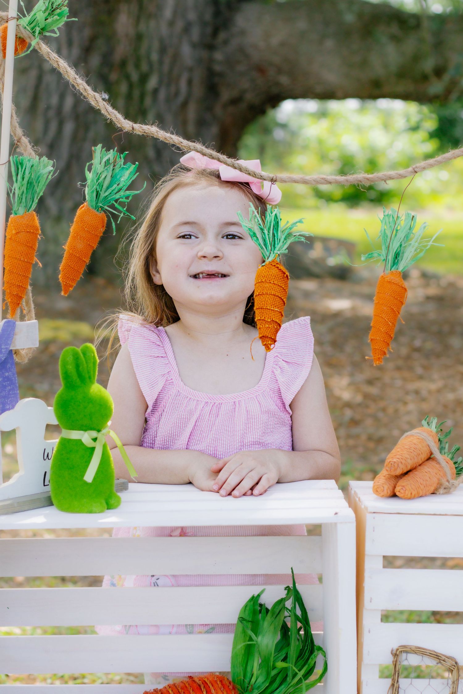 Smiling girl in pink dress poses at Easter display with carrot garland and bunny decorations.