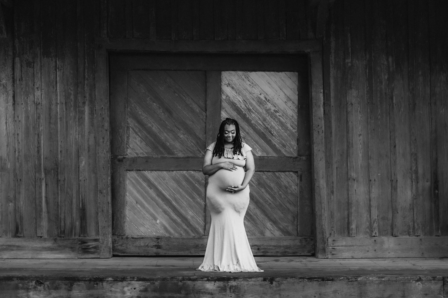 Black and white maternity photo of pregnant woman in flowing gown posing before wooden barn doors.