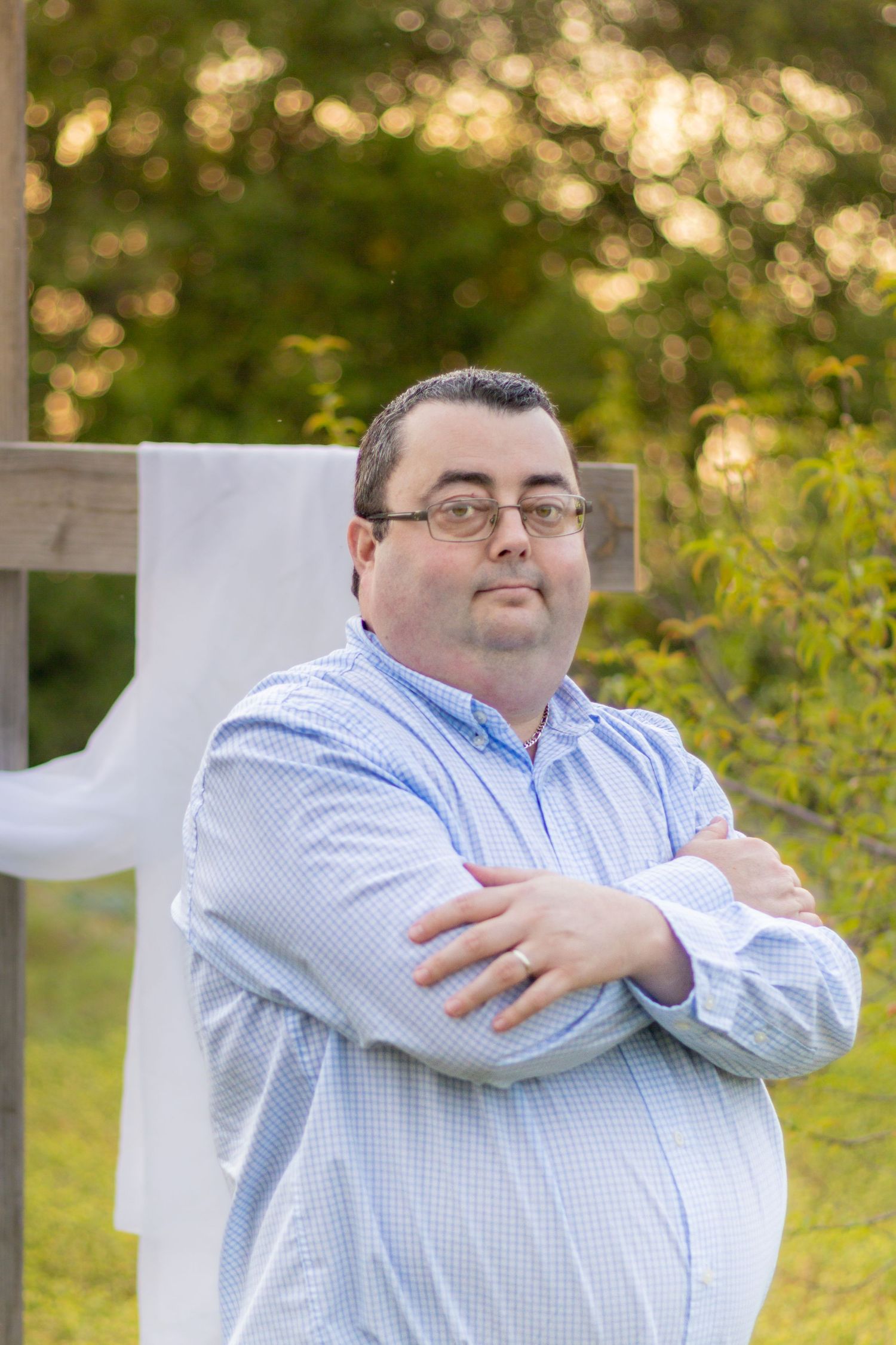 Man with glasses in light blue shirt standing with arms crossed near a white-draped wooden cross outdoors.
