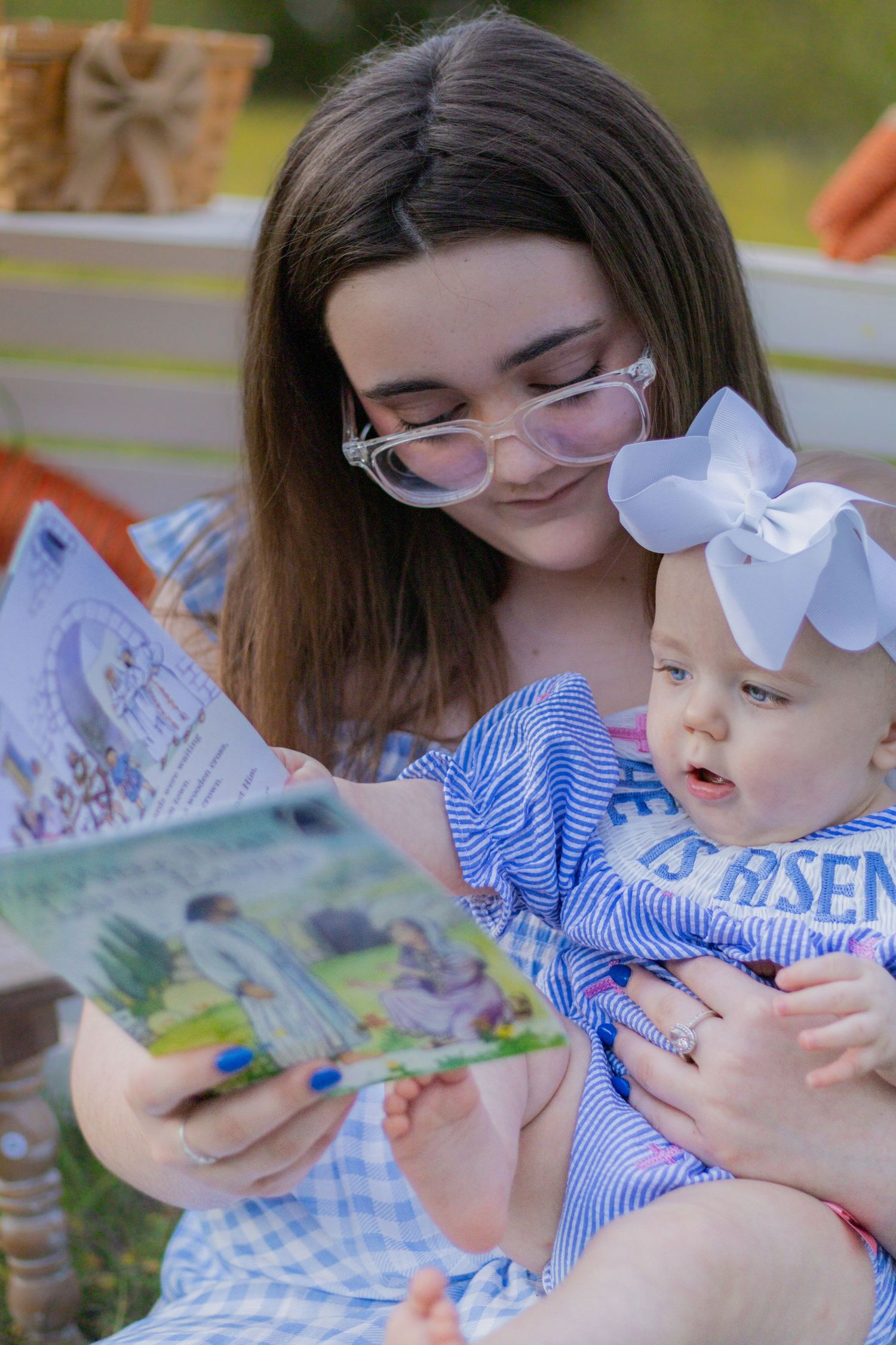 Young woman with glasses reads Easter book to baby with white bow headband outdoors.