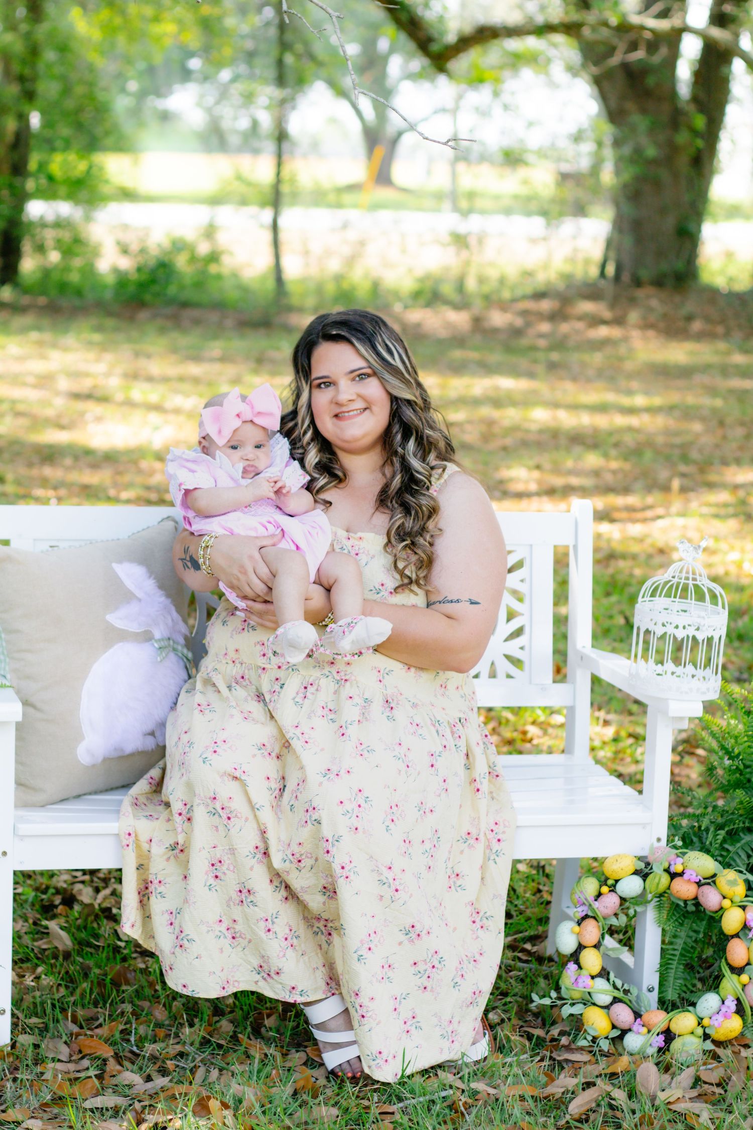 Smiling mother in yellow floral dress holds pink-dressed baby on white bench with Easter decorations.