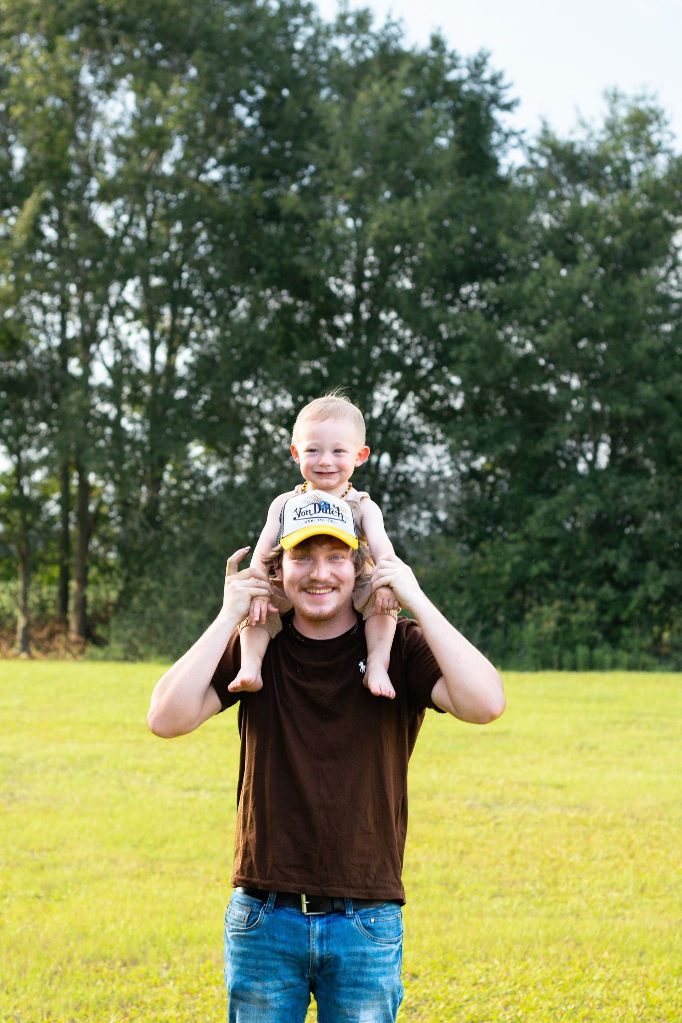 A smiling man in a brown shirt holds a happy toddler on his shoulders outdoors on a green lawn.