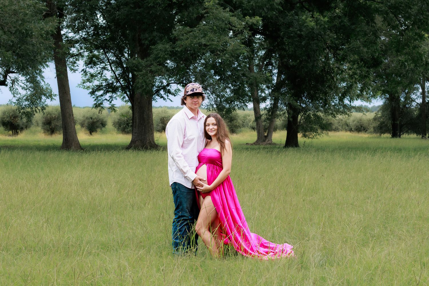 Couple posing in green field, woman in pink maternity gown, man in white shirt and cap, trees in background.