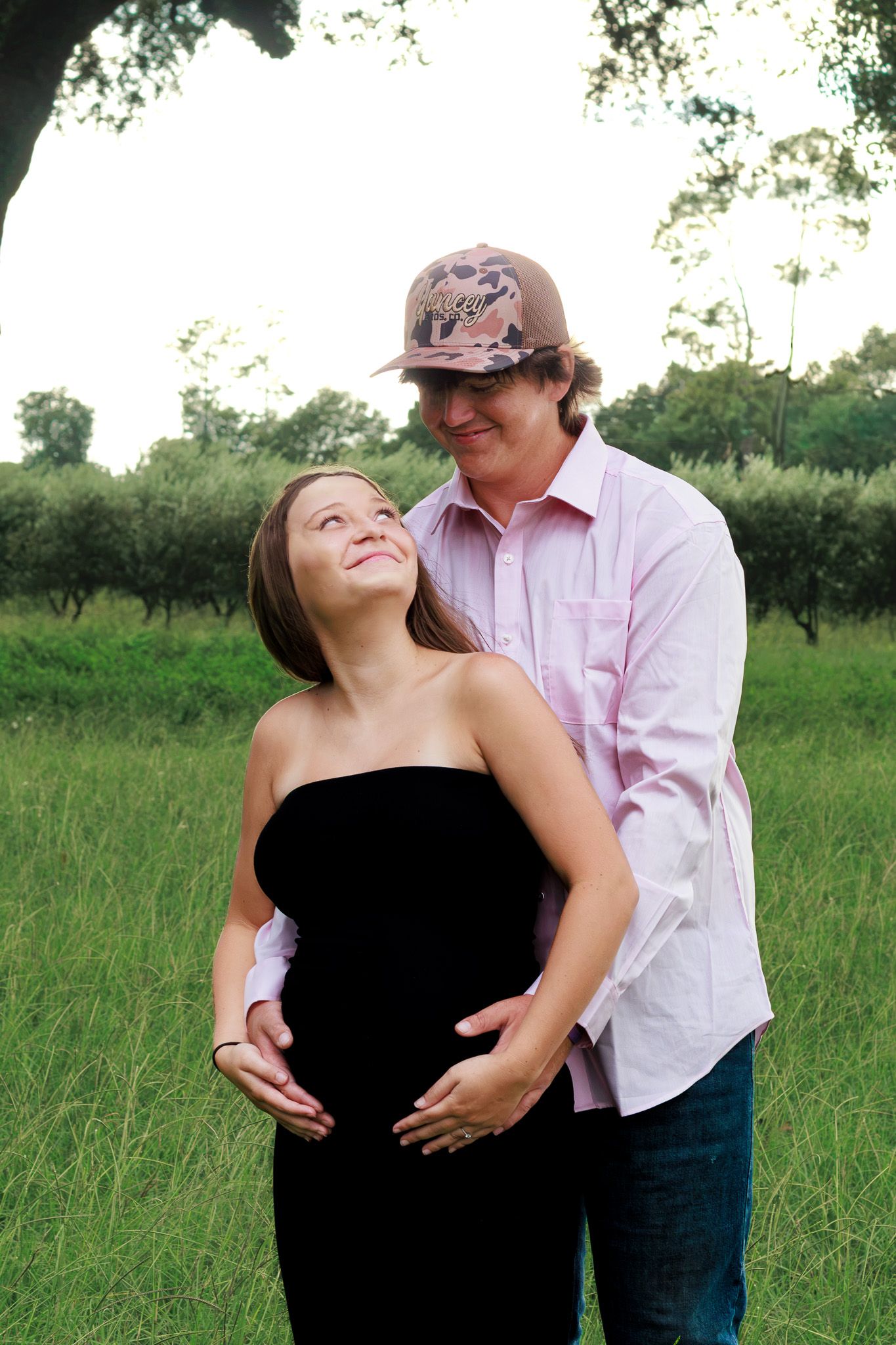 Pregnant couple embracing outdoors in green field, man in pink shirt and camo hat, woman in black strapless dress.
