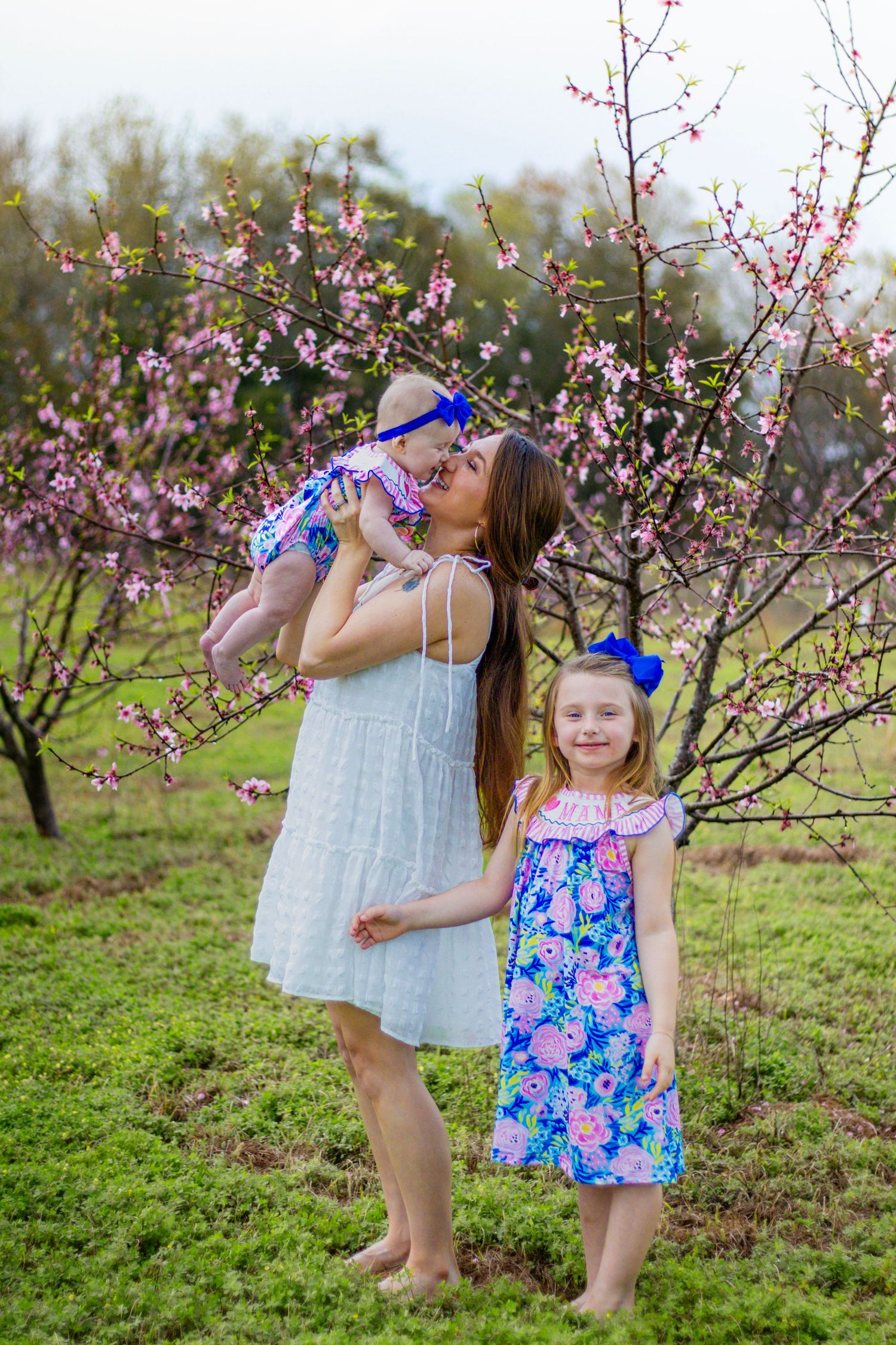 Mother in white dress kissing baby while young girl in floral dress stands beside her among pink blooming trees.