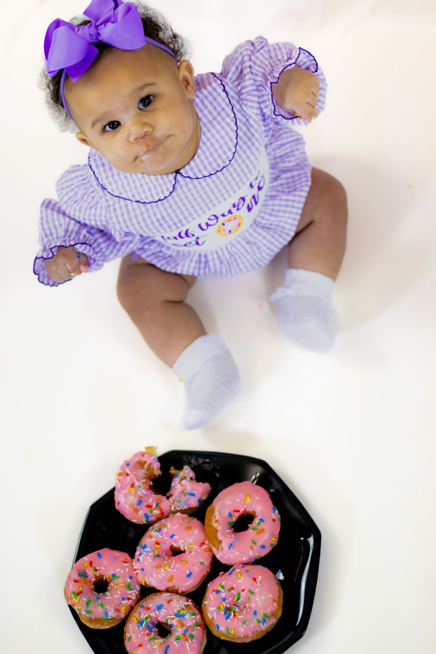 Overhead view of baby girl in purple outfit beside plate of pink sprinkle donuts.