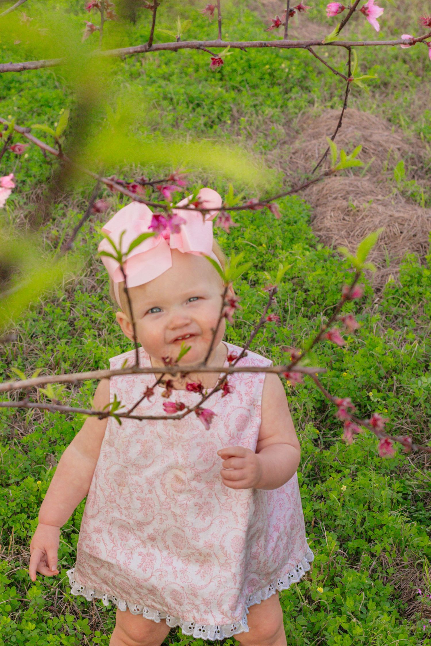 Smiling baby girl in pink lace dress with bow headband standing among blooming spring cherry blossom branches.