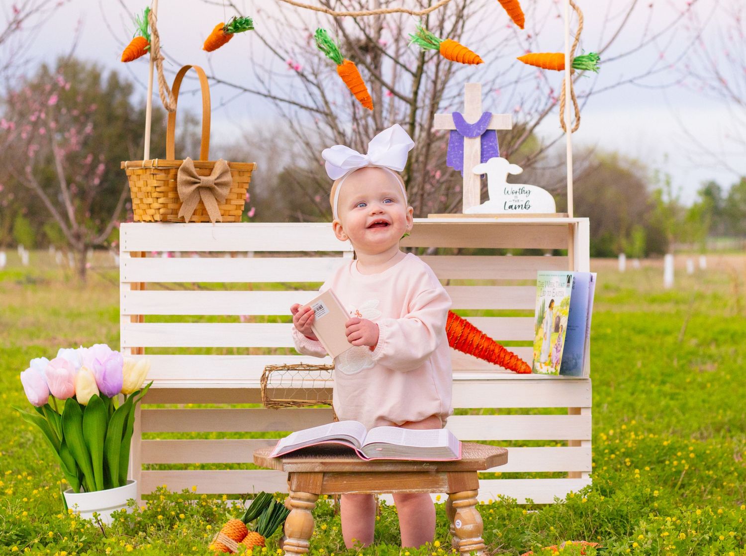 Laughing baby with bunny ears sits on bench with open book at outdoor Easter display.
