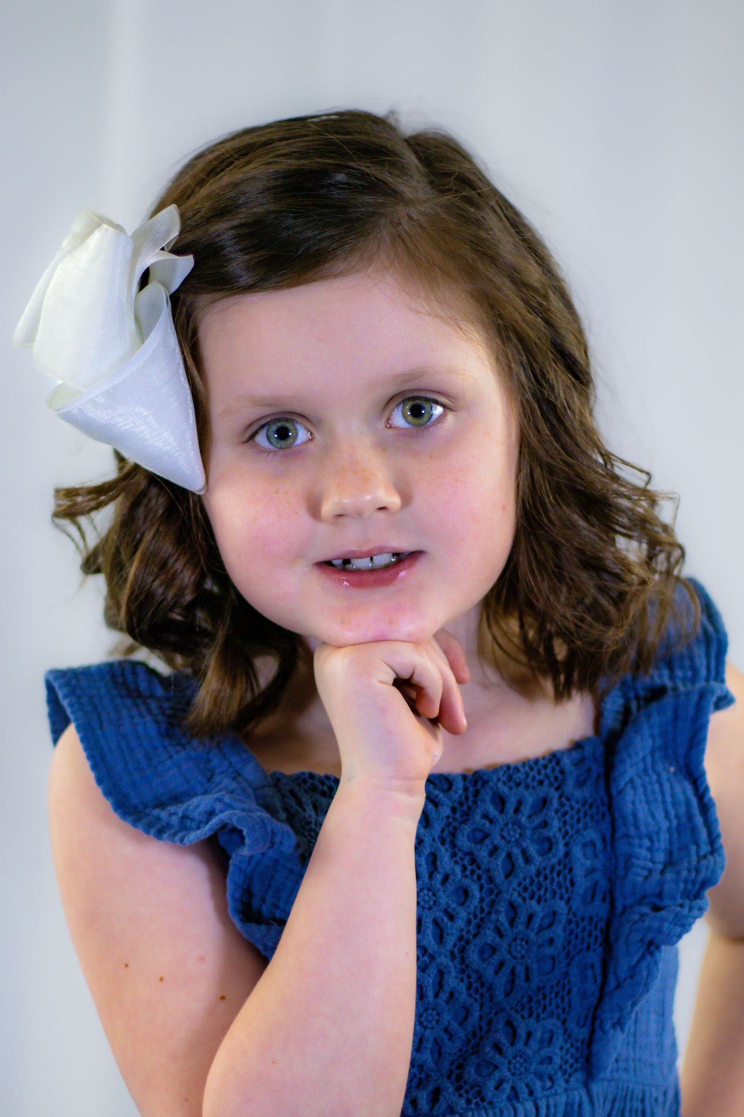 Young girl in blue dress with white bow poses thoughtfully with chin resting on hand.