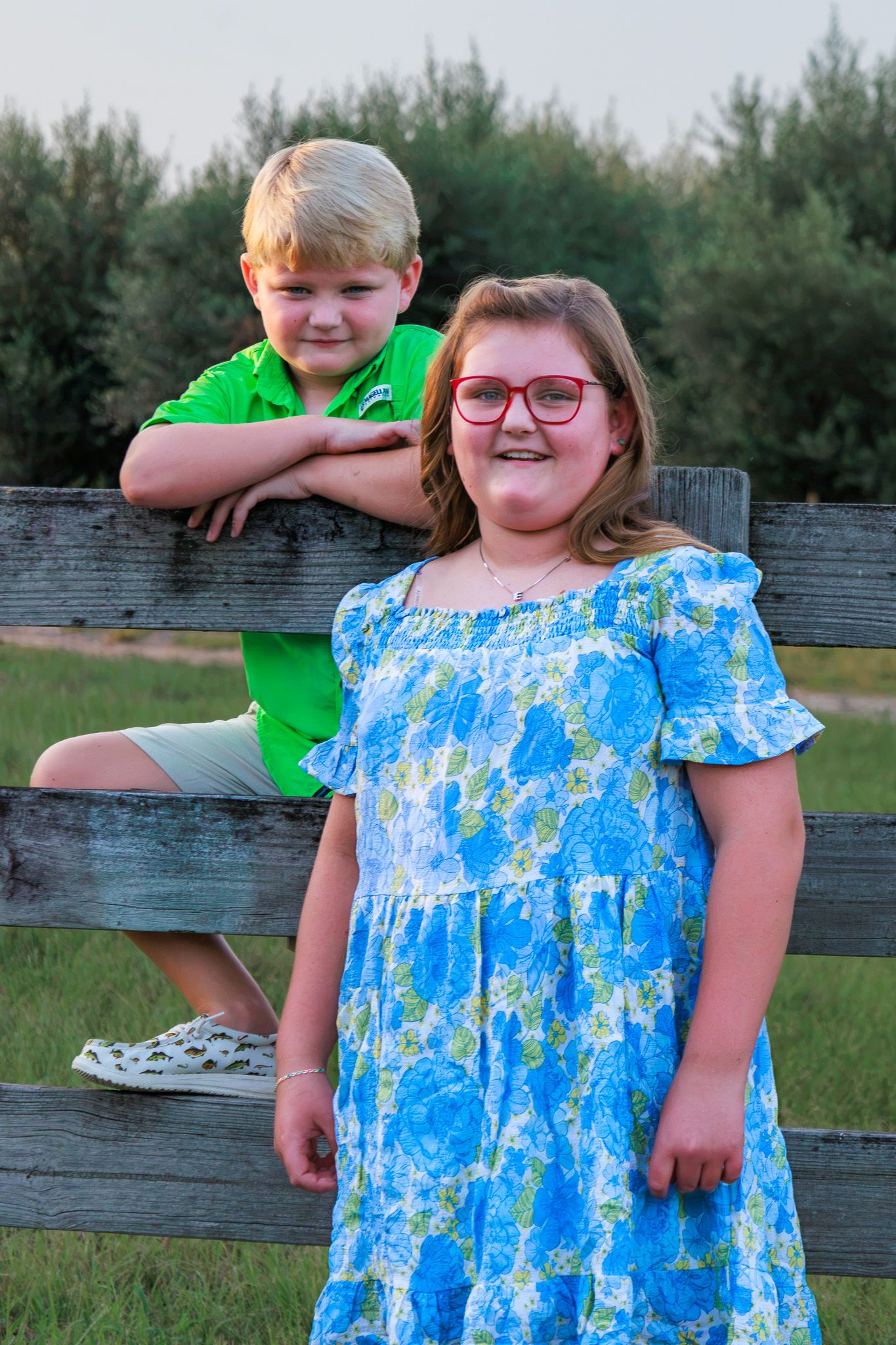 Two children pose by a rustic wooden fence outdoors, a boy in green polo and a girl in blue floral dress wearing red glasses.
