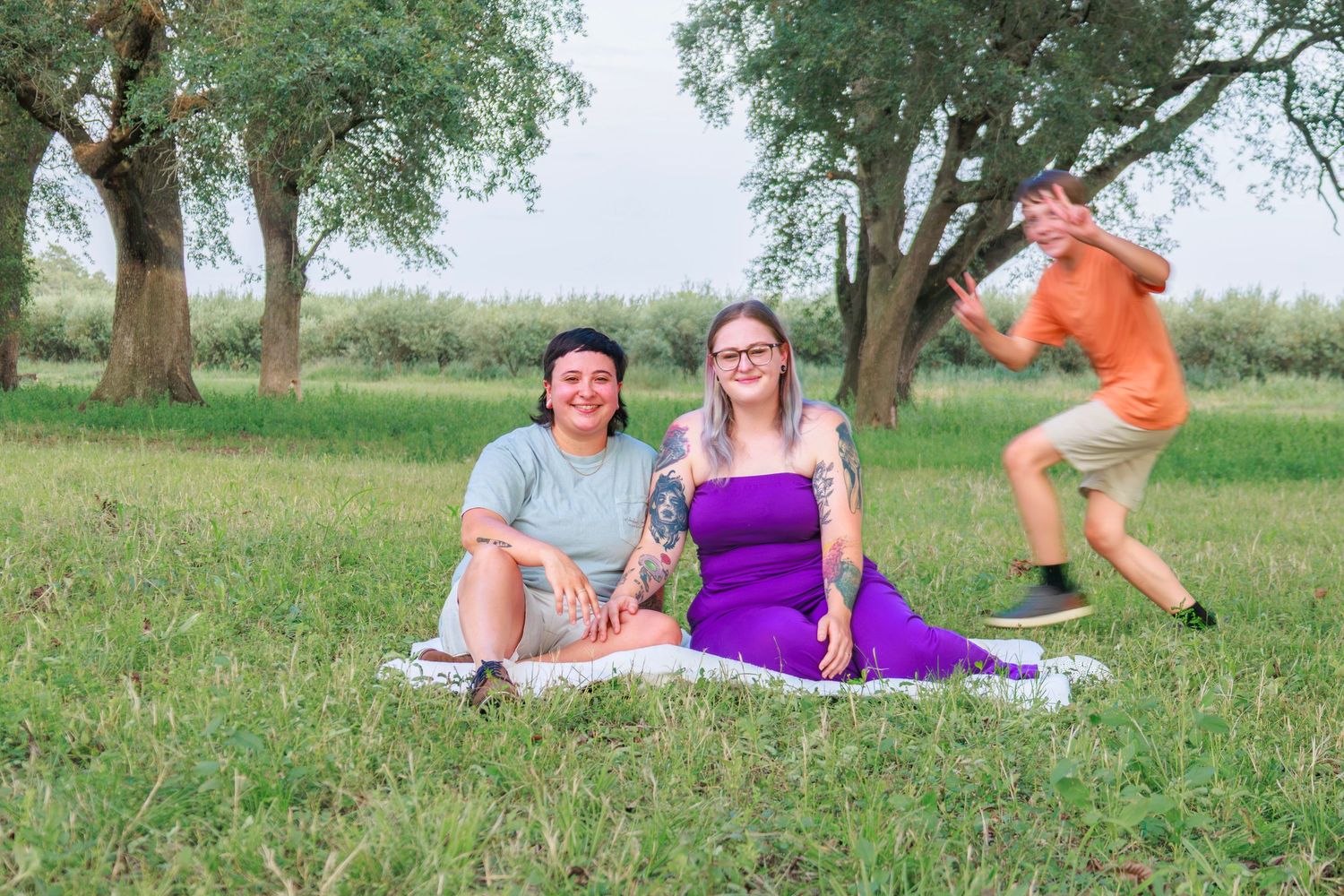 Two people sit on a blanket in a green park while a person photobombs in the background.