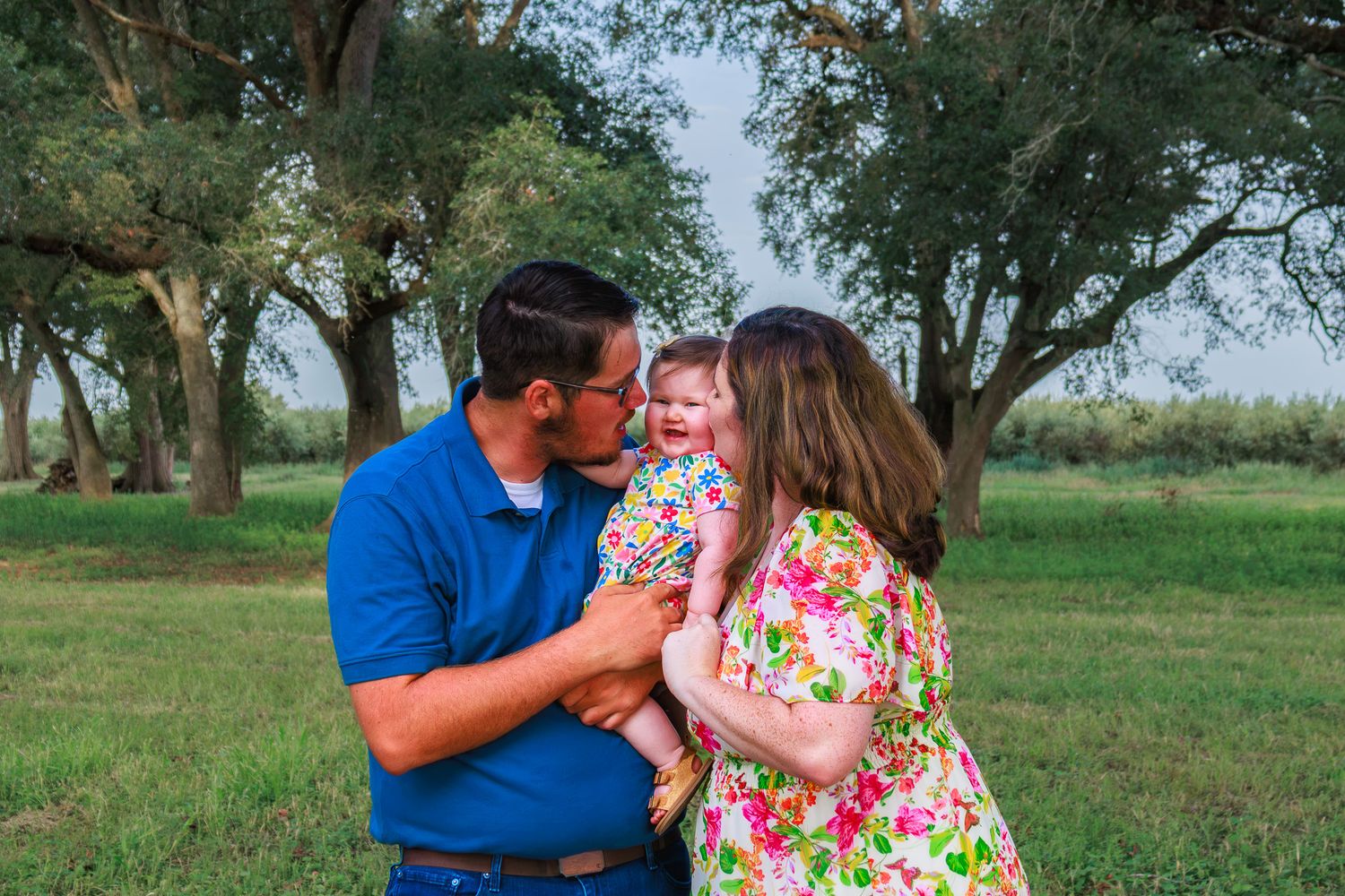 A couple kisses their smiling baby girl outdoors in a lush green field with large trees in the background.