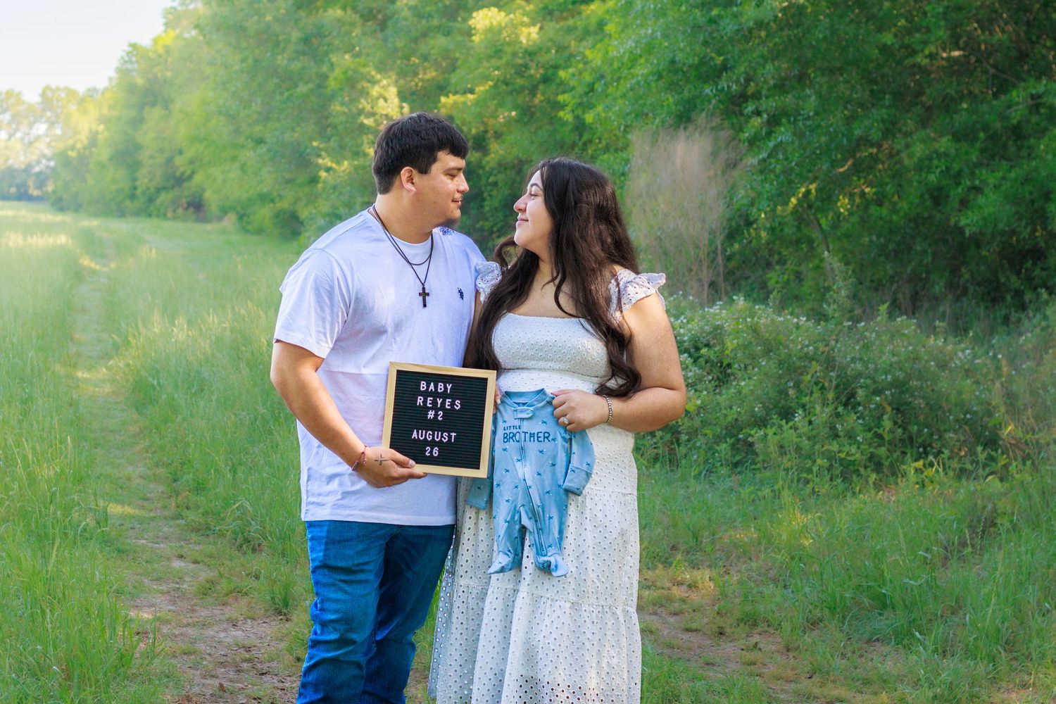 Expecting couple holds Baby Reyes announcement board and blue outfit on scenic green path.