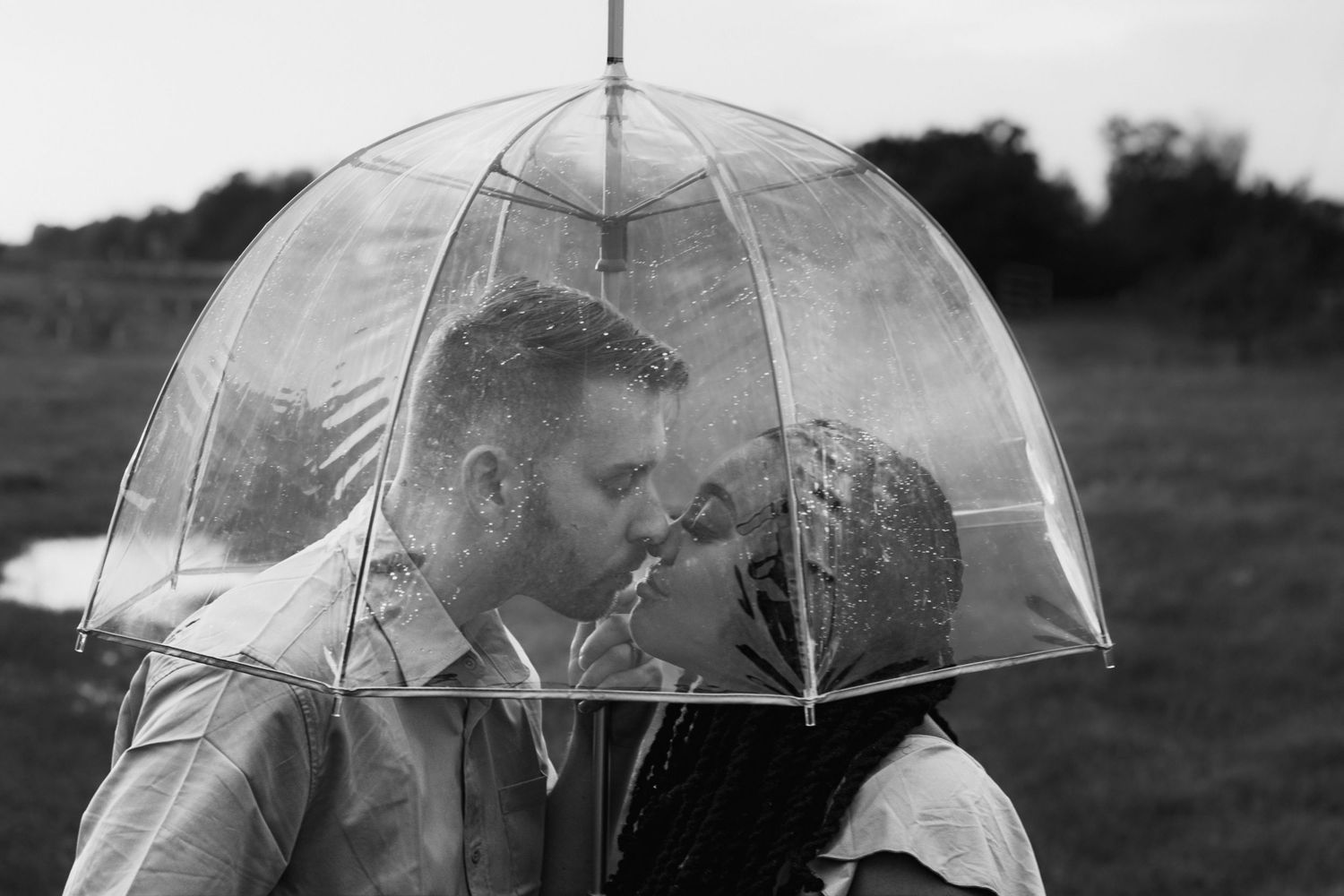 Couple shares romantic kiss under clear bubble umbrella in black and white outdoor maternity photo.