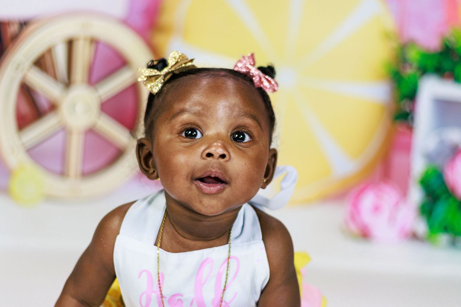 Cute baby girl with gold and pink bows smiling in front of a yellow lemon-themed birthday photo backdrop.